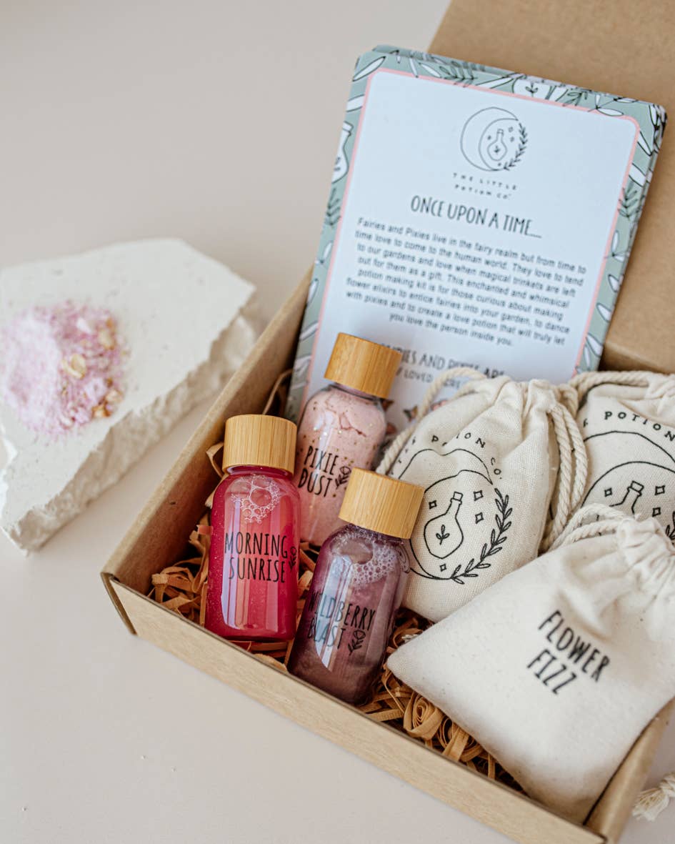 The Enchanted Garden MINDFUL Potion Kit by THE LITTLE POTION CO includes small bottles labeled 'Morning Sunrise,' 'Pixie Dust,' and 'Blueberry Potion.' It also features cloth bags labeled 'Potion' and 'Flower Fizz,' along with a note titled 'Once Upon a Time' that boasts a whimsical design. Completing the mindful magic collection, there's a heart-shaped bath bomb nearby.