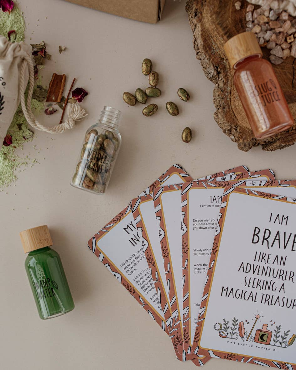 A flat lay image showcasing the Wild Adventure MINDFUL Potion Kit by THE LITTLE POTION CO, featuring cards with motivational quotes, small bottles of colorful liquids reminiscent of a potion kit, and scattered seeds and herbs. One card reads "I am brave like an adventurer seeking a magical treasure," while natural elements and rustic items add an earthy tone.