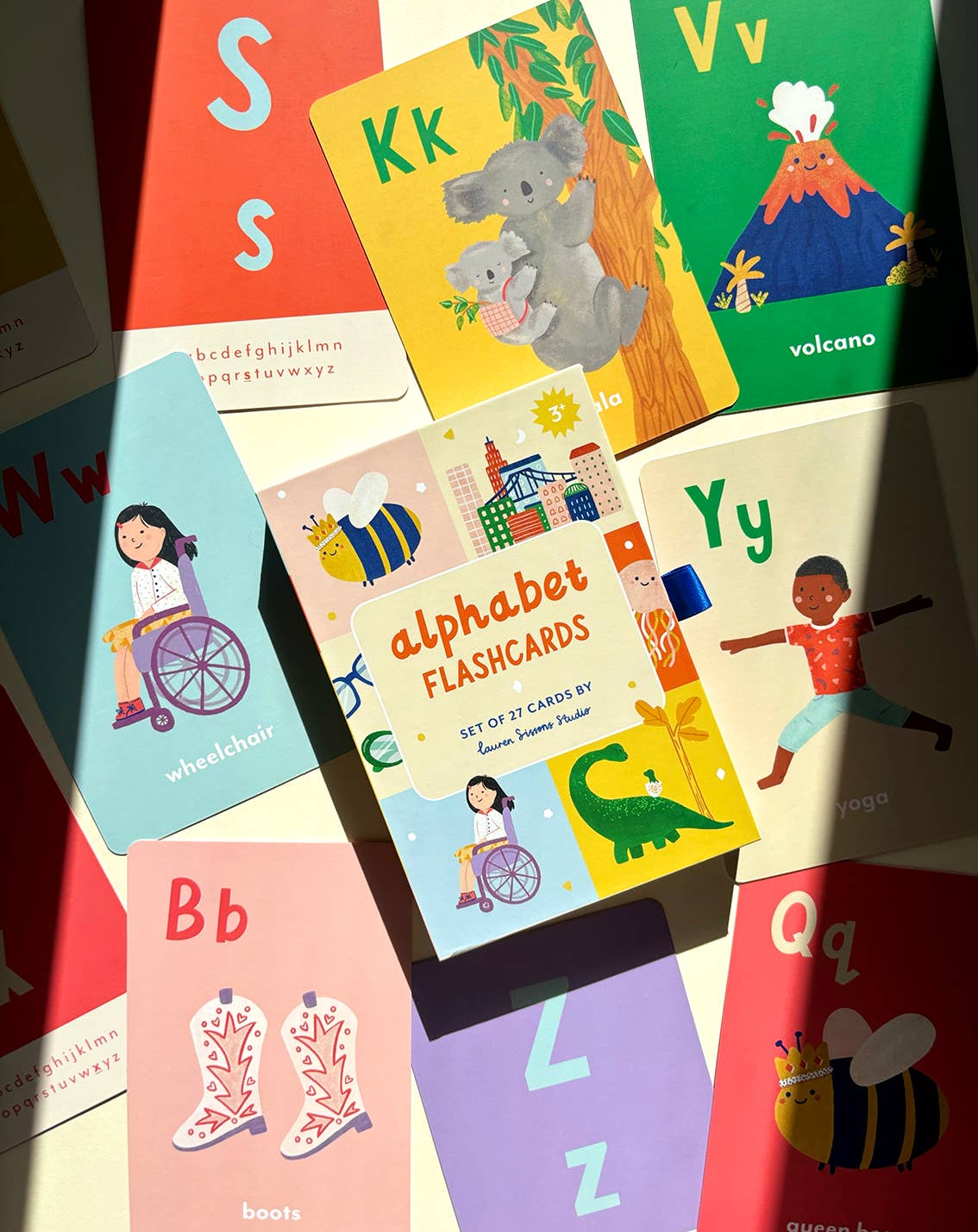 A collection of vibrant, laminated alphabet flashcards is spread out on a surface, surrounding a box labeled "A-Z Illustrated Alphabet Flashcards" by LAUREN SISSONS STUDIO. Each card showcases a letter accompanied by an illustration, such as "W" for wheelchair, "V" for volcano, and "Y" for yoga. Sunlight casts a shadow across these children's educational toys.