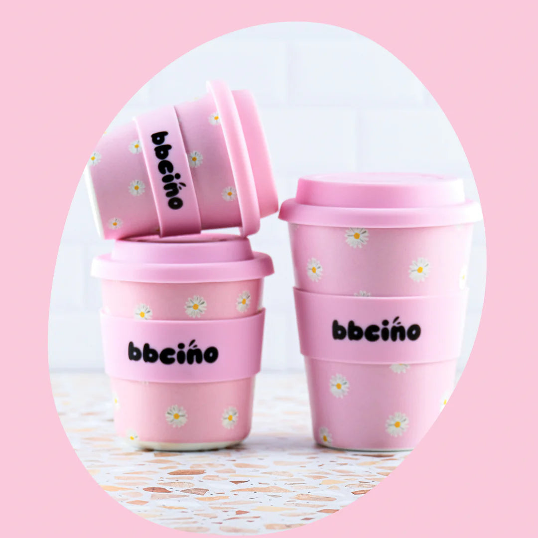 A set of four BBCINO Reusable BambinoCino Cups (240ml) in Daisy Baby Pink, featuring double size capacity and complemented by silicone straws. The cups are arranged in pairs on a pastel pink background, some positioned against a white tiled wall and terrazzo surface. Two cups stand upright while the other two are tilted, leaning on each other.