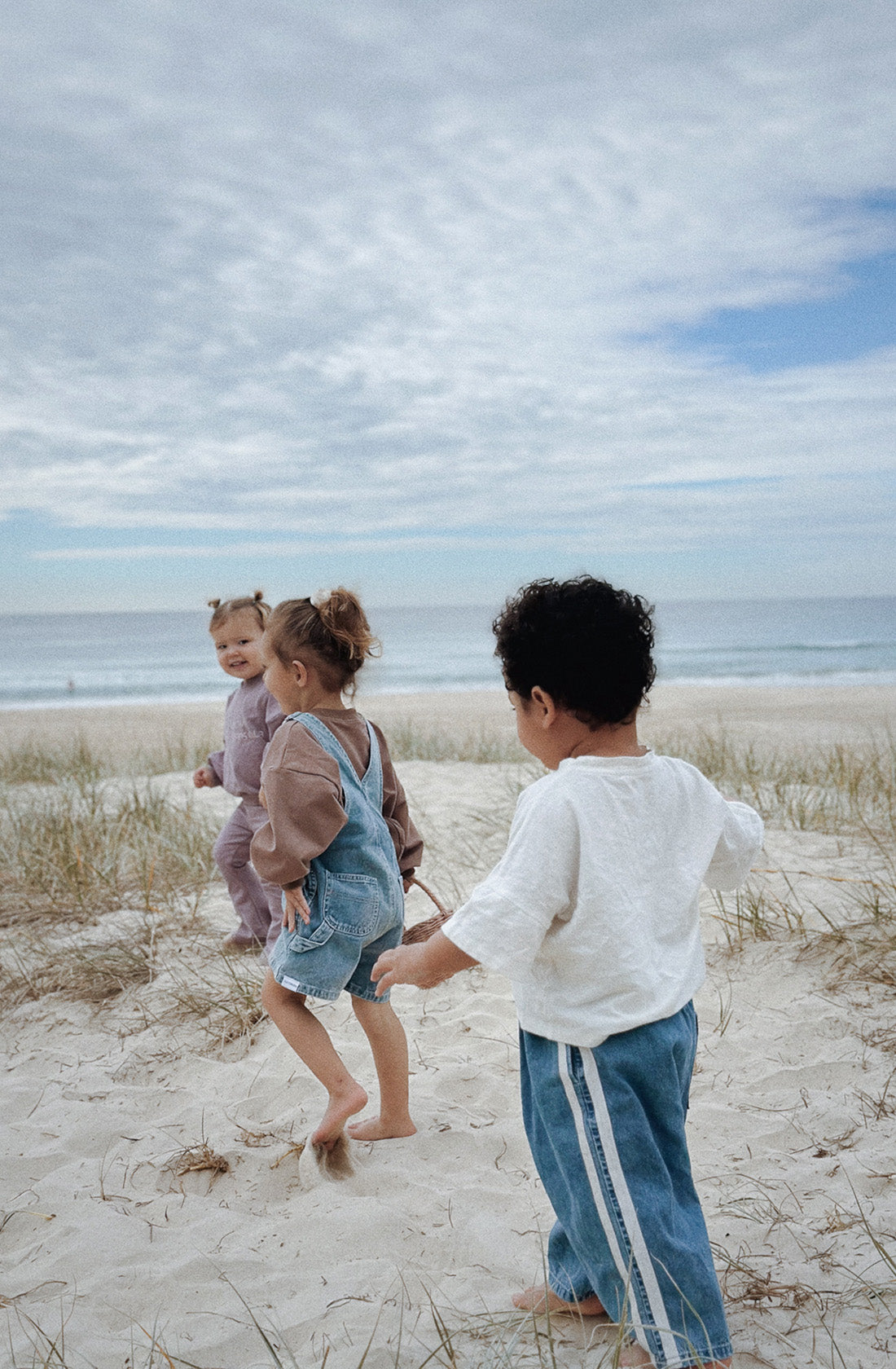 Three young children are playing on a sandy beach with grass patches. Two children, wearing TWIN COLLECTIVE Tricky Track Pants, hold hands and run while the third child follows behind. The sky is partly cloudy, and the ocean is visible in the background. The children appear joyful and playful.