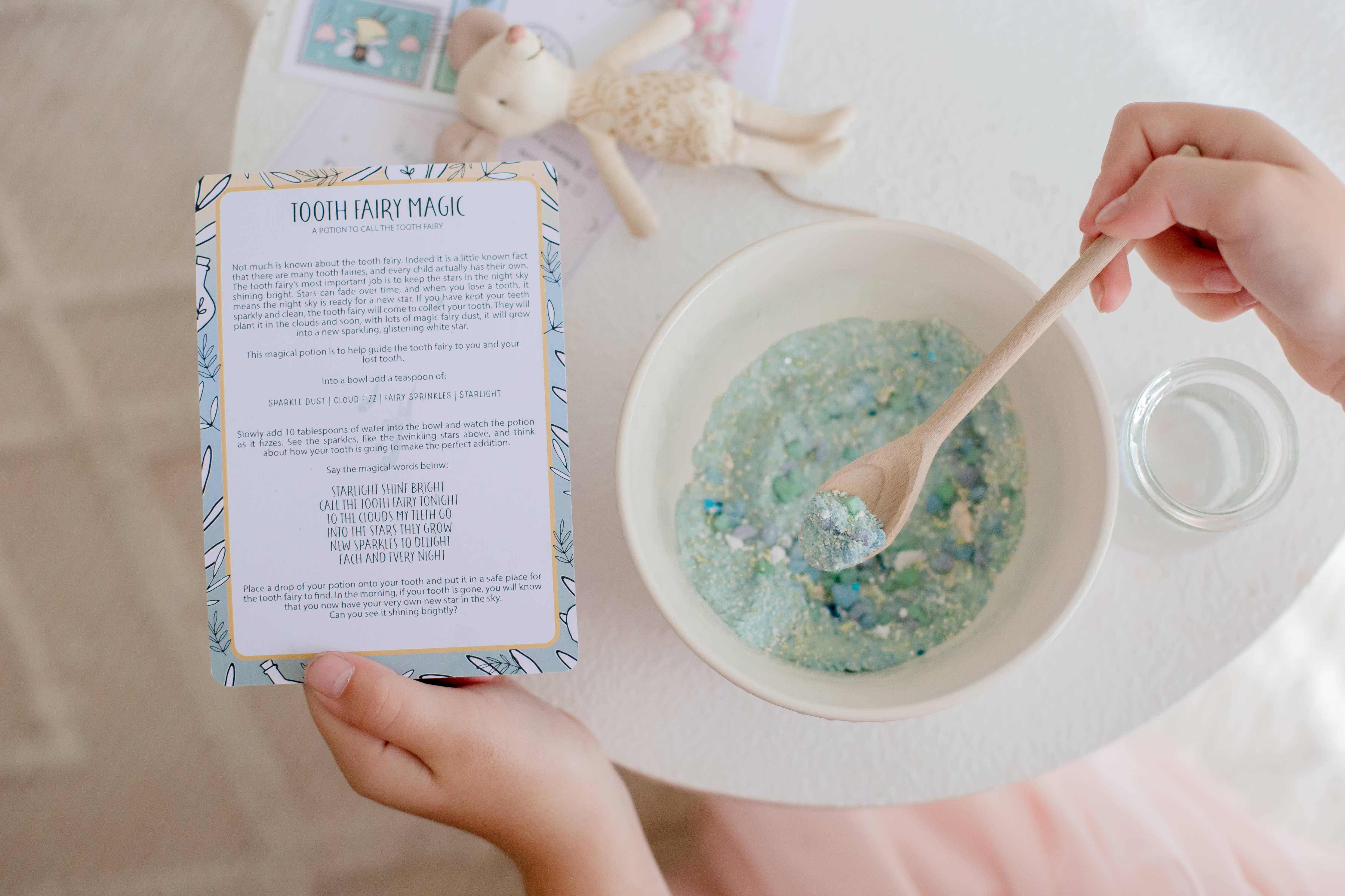 A child holds a package labeled "Tooth Fairy Magic Potion Pouch" from THE LITTLE POTION CO next to a bowl containing blue and green sparkling powder, reminiscent of secret tooth fairy potions. A wooden spoon sits in the bowl, while a small glass jar and a toy rabbit are nearby on the table.