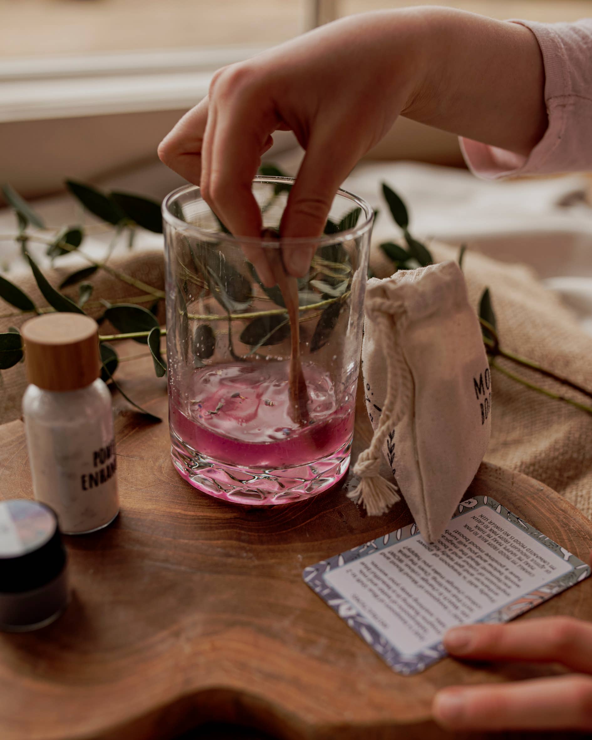 A hand stirs a pink liquid in a glass with a wooden stick from THE LITTLE POTION CO's MINI Colour Mood Potion Kit (Colour Changing Kit), placed on a wooden board. Nearby items include a small bottle, a pouch, and a card with text. The background features green leaves and neutral-colored fabric, evoking the enchanting vibe of magic potion kits.