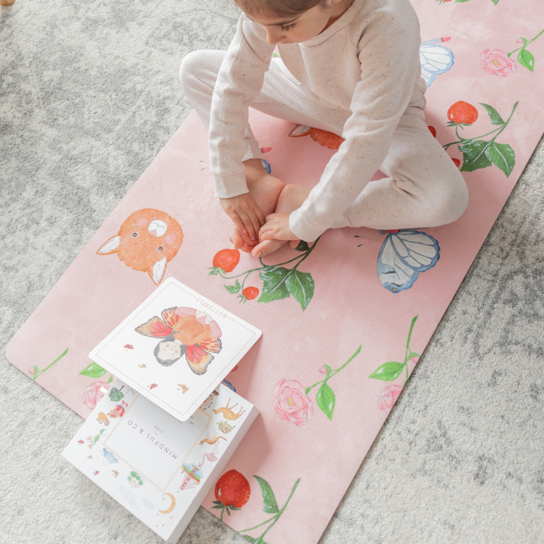 A young girl sitting in a meditation pose on the seet themed pink kids yoga mat with illustrated cards.