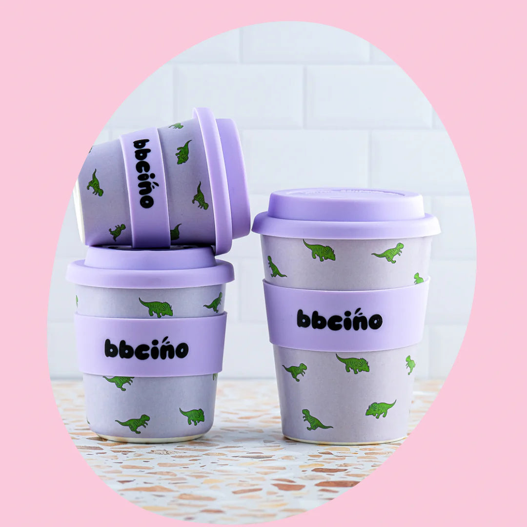 Three stacked Reusable BambinoCino Cups 240ml Dino-mite by BBCINO, adorned with purple lids and bands, green dinosaur designs, and the text "BambinoCino reusable cup." The background features a pink hue with a white tile pattern. The cups are displayed on a terrazzo-style surface and each comes with a reusable silicone straw.