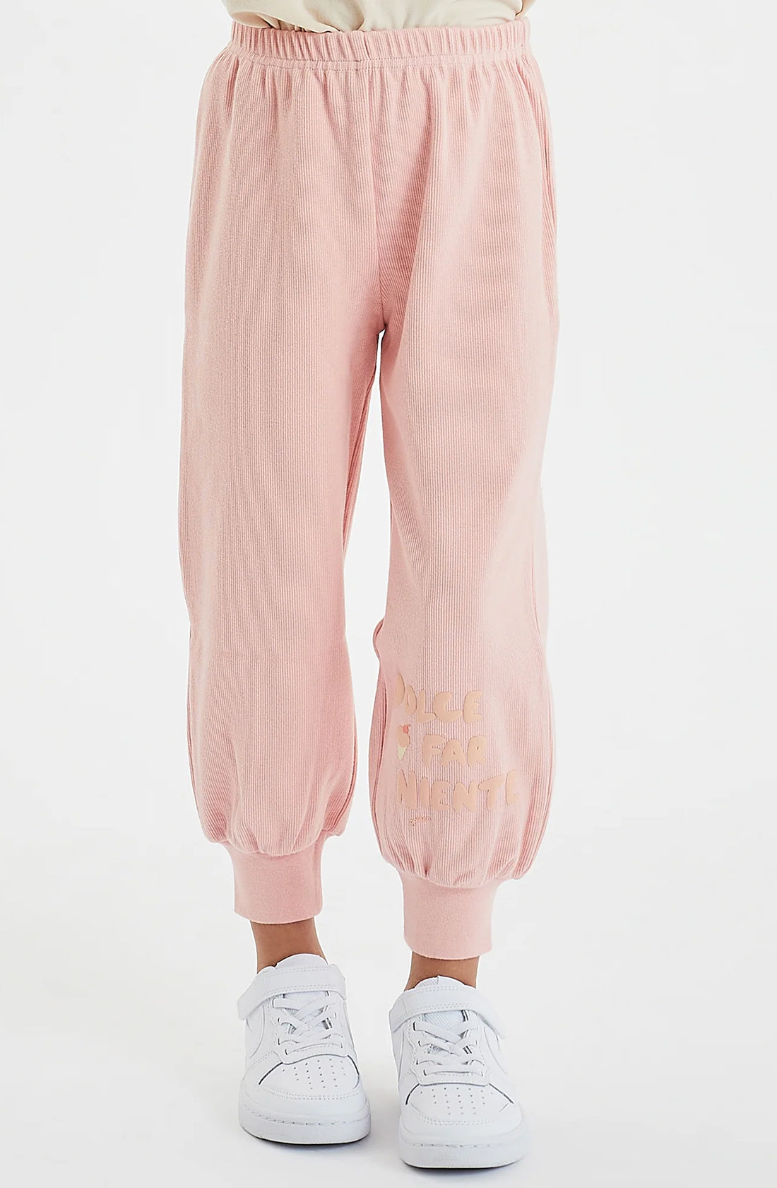 An individual is standing in a light-colored room wearing GOLDEN CHILDREN's Dolce Lounge Pant in Pink Lemonade, which includes an elastic waistband and cuffs. These 100% cotton rib joggers feature some embroidered text near the bottom on one leg. The person is also wearing white sneakers, with their upper body not visible.