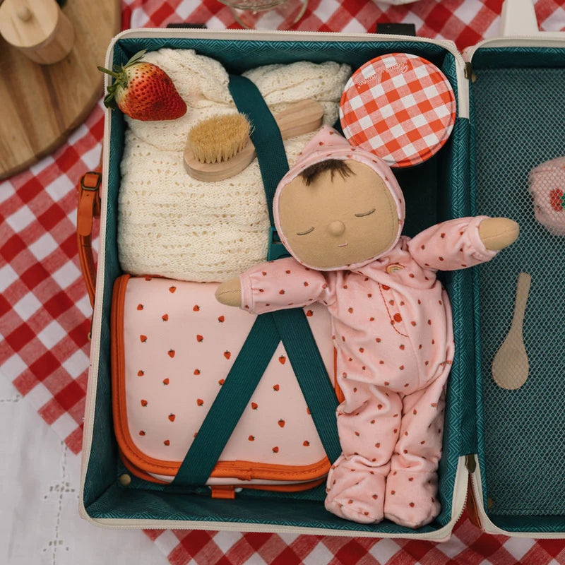 A small suitcase contains a Dozy Dinkum Strawberry Pip doll in pink pajamas, a cream knitted garment, a strawberry, a small brush, an Olli Ella jar with a red and white checkered lid, and other small items. The suitcase is lined and displayed on a red and white checkered tablecloth.