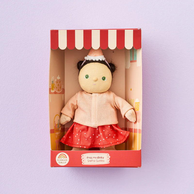 The Dress Me Dinky Sophia Sundae doll by OLLI ELLA features a round face with black hair styled in buns, wearing a light pink jacket and a red skirt with white dots. Nestled in a box with charming dessert illustrations and a rainbow, it showcases a red and white striped top designed for creative play.