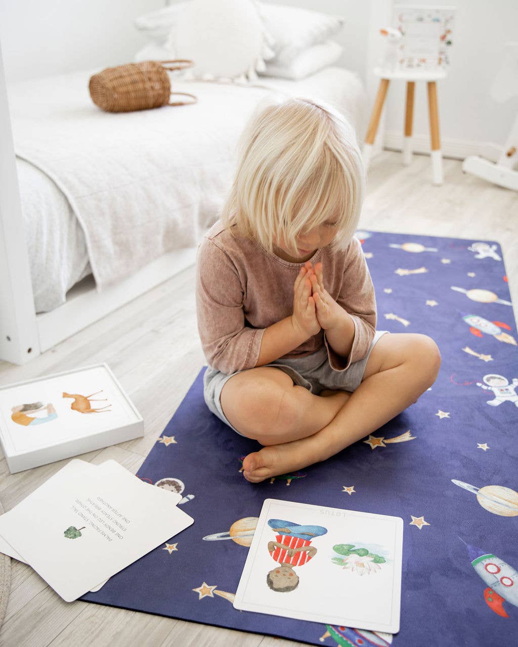 A young kid sitting on a navy blue yoga mat with books arounf him with his hands in the pray position.