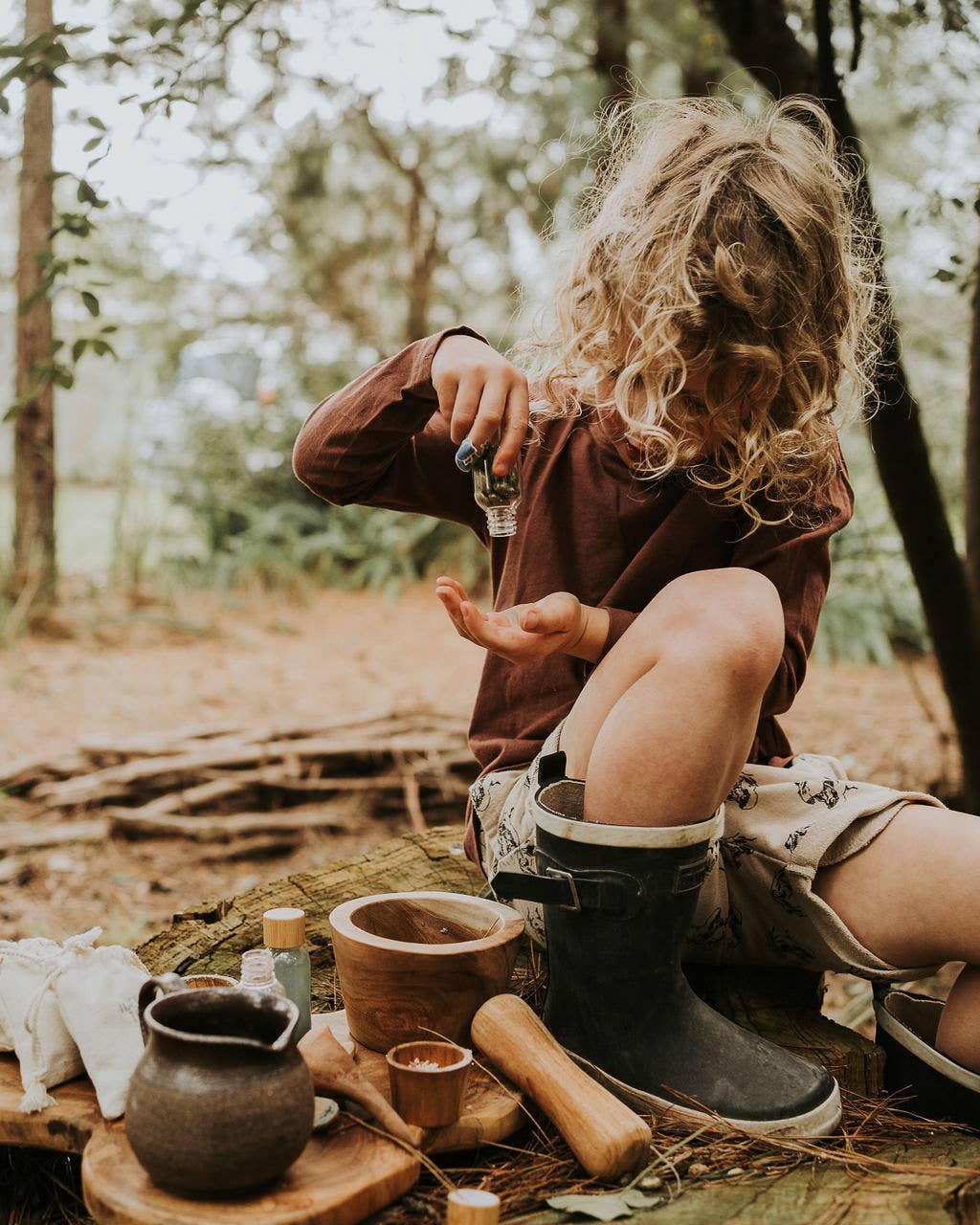 A child with curly hair, wearing a brown shirt, shorts, and black rubber boots, is sitting on a tree stump in a forest. The child is using the Wild Adventure MINDFUL Potion Kit by THE LITTLE POTION CO, meticulously pouring liquid from a small bottle into their hand as though mixing magic potions. They are surrounded by wooden bowls and natural materials on the stump.