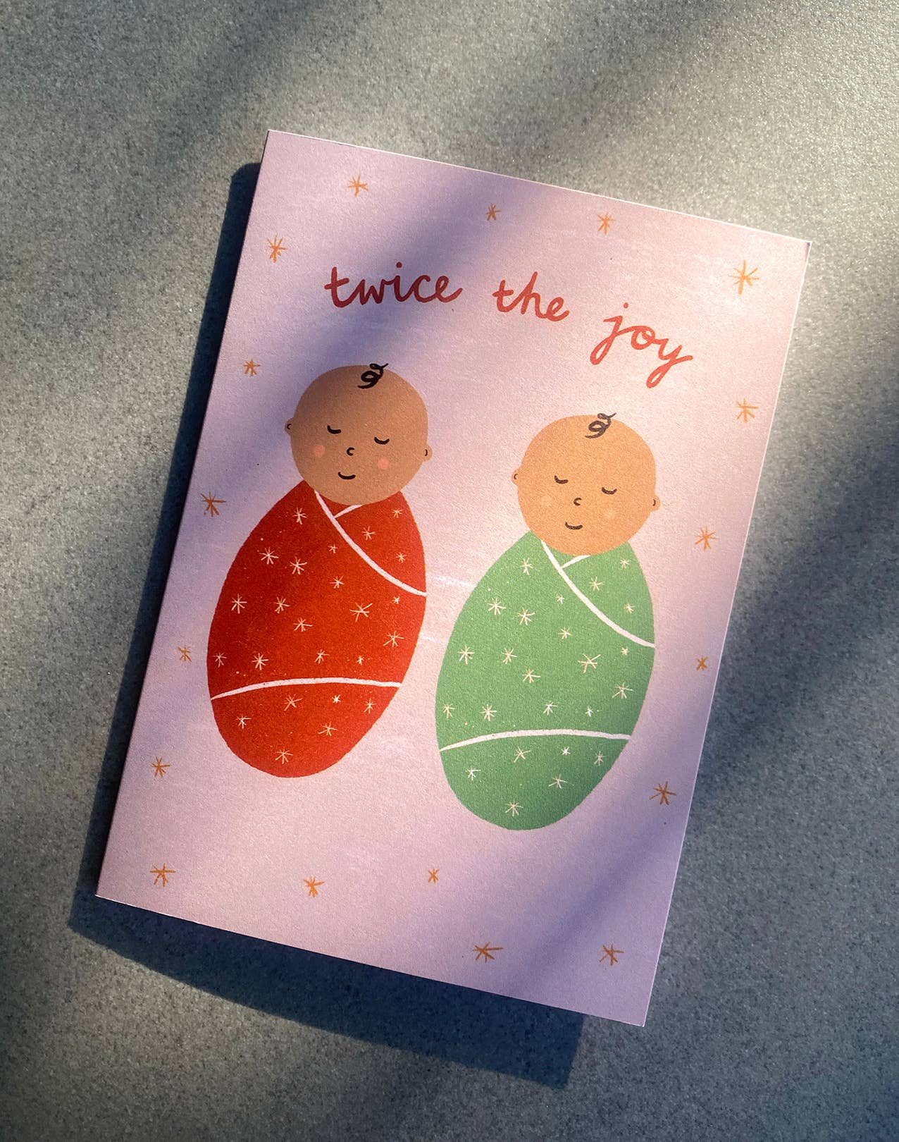 The "Twice The Joy" New Baby Greeting Card by LAUREN SISSONS STUDIO features two illustrated babies swaddled in blankets—one in red and the other in green. Handwritten text in orange above them reads "twice the joy." Perfect for celebrating new baby twins, this card is set against a simple background with a soft shadow and is printed on recycled paper.