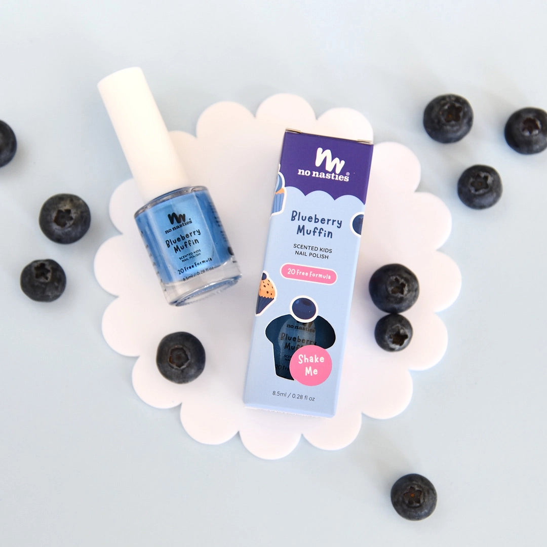 The NO NASTIES Scented Scratch Off Kids Nail Polish in Blueberry Muffin/Blue sits on a white flower-shaped mat, surrounded by blueberries, with its playful, branded box displayed beside it.