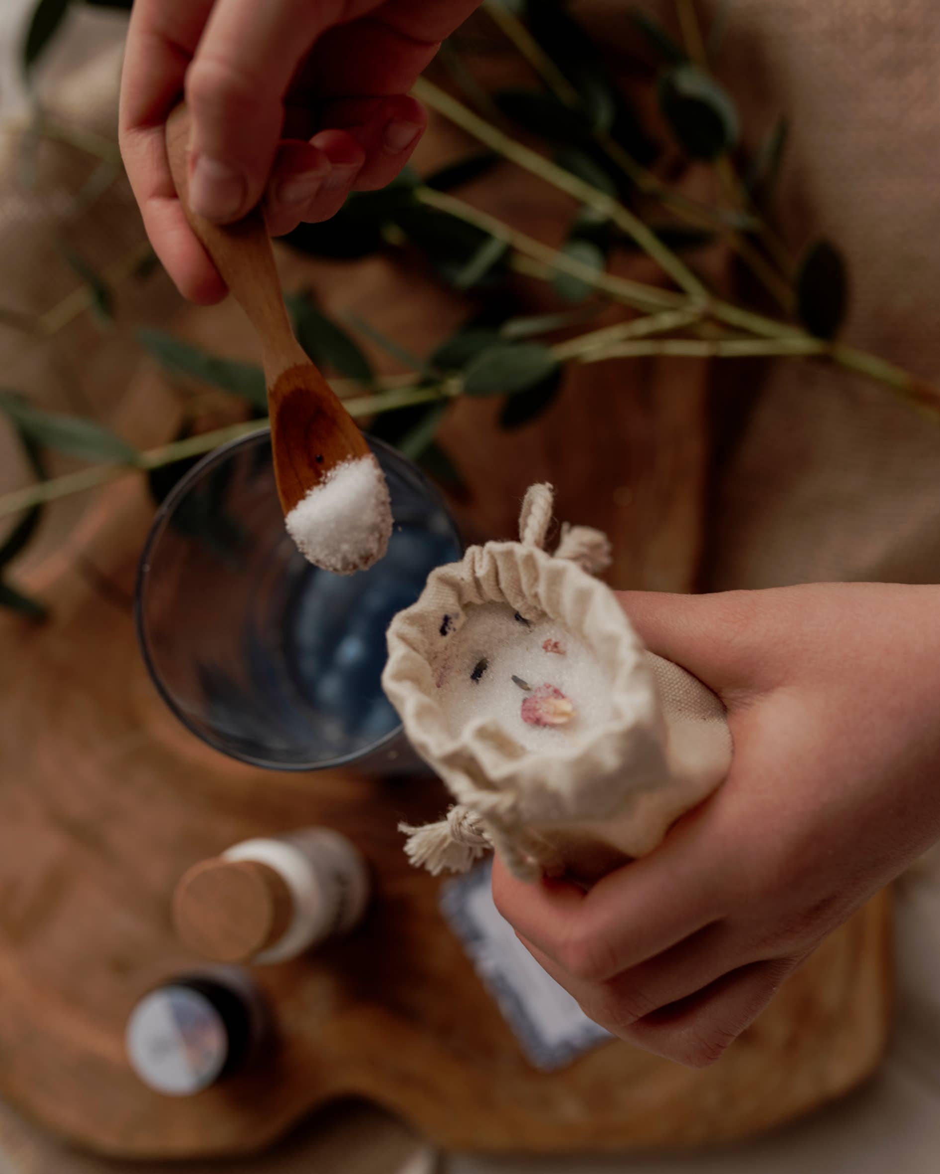 A hand holding a small wooden spoon of white powder above a glass, with another hand holding a drawstring cloth bag from THE LITTLE POTION CO's MINI Colour Mood Potion Kit (Colour Changing Kit). Green leaves and small bottles are scattered on the wooden surface in the background, adding an air of mystique to the scene.