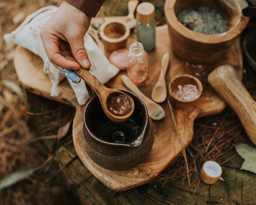 A hand is holding a wooden spoon with a brown substance over a ceramic bowl. The table, resembling the Wild Adventure MINDFUL Potion Kit by THE LITTLE POTION CO, is filled with various items including bowls, wooden utensils, a bag, and bottles—all arranged on a rustic wooden surface outdoors. The setting has a natural feel that invites mindfulness.