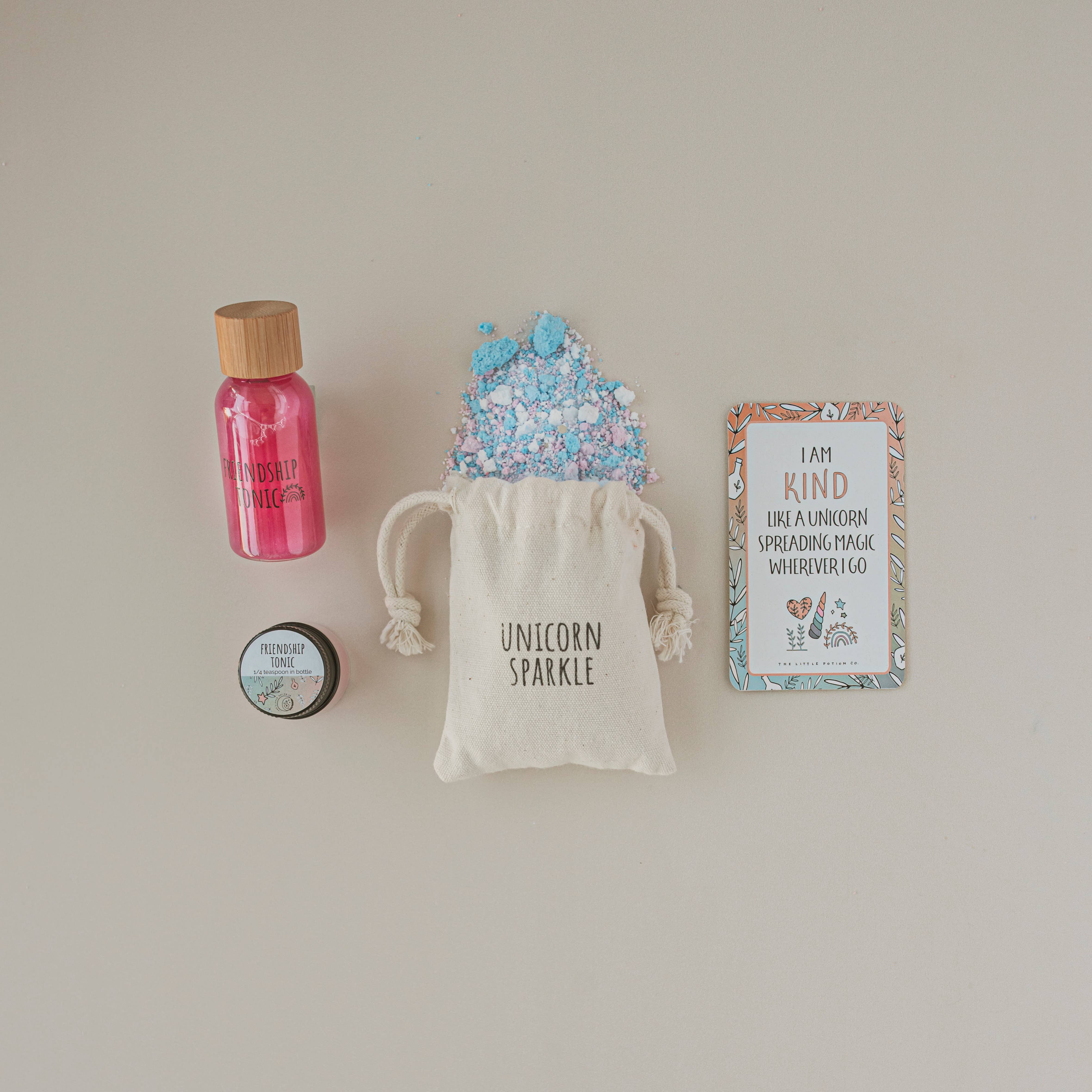 A flat lay image showcases items for imaginative play from THE LITTLE POTION CO's MINI Rainbow Sparkles Potion Kit: a small glass bottle filled with pink liquid labeled "Unicorn Magic," a sealed jar labeled "Fairy Grove Mushroom Lip Balm," a beige drawstring bag labeled "Unicorn Sparkle" with blue and pink sparkles spilling out, and a colorful card that reads "I AM KIND LIKE A UNICORN SPREADING MAGIC WHEREVER I GO.