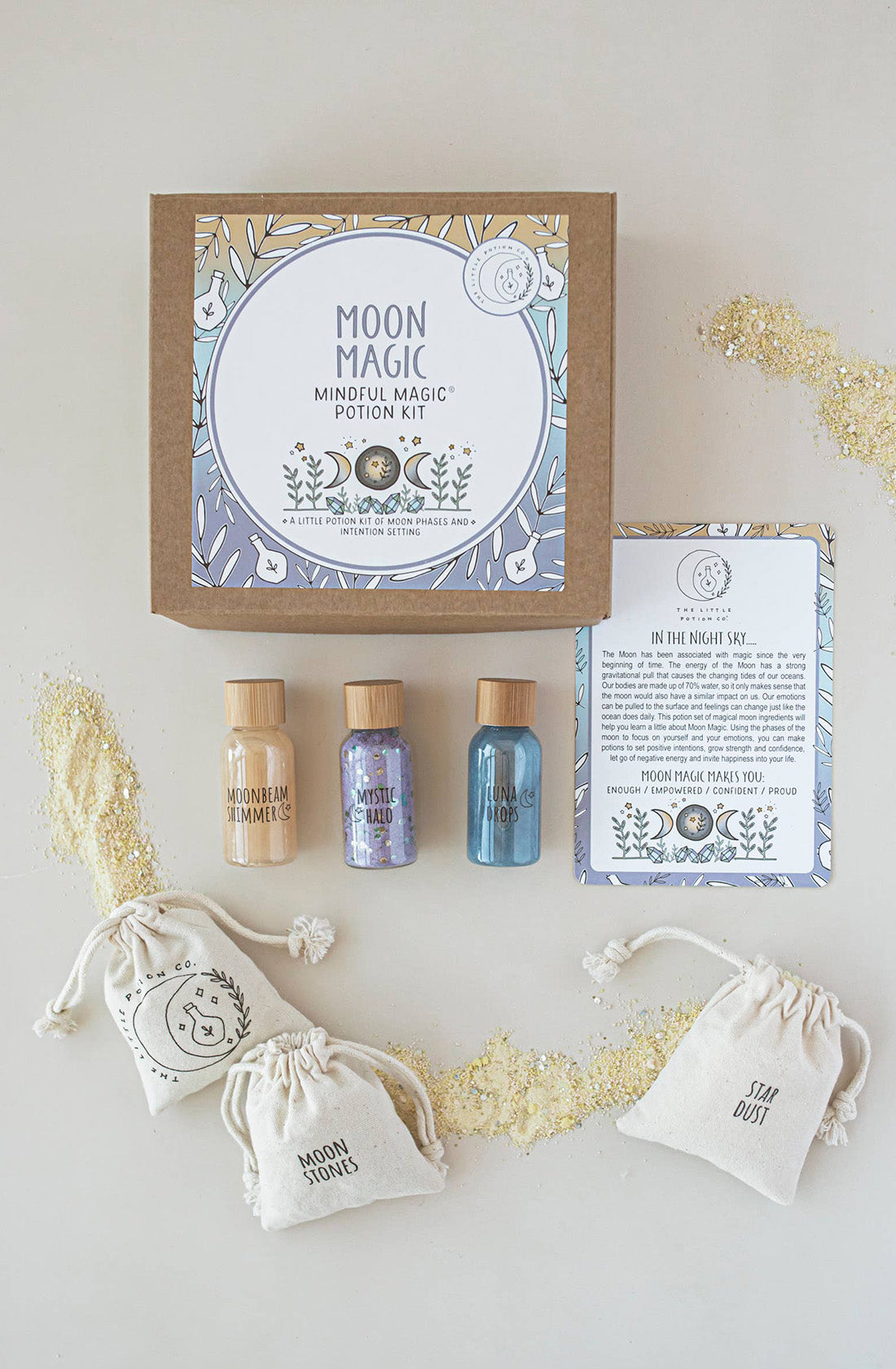 A Moon Magic MINDFUL Magic Potion Kit from THE LITTLE POTION CO is displayed, including a box, three potion bottles labeled Moonbeam Glimmers, Mystic Haze, and Luna Glow. It also features two drawstring pouches labeled Moon Stones and Star Dust, along with an instruction sheet detailing a mindfulness activity for each moon phase on a light background with scattered glitter.