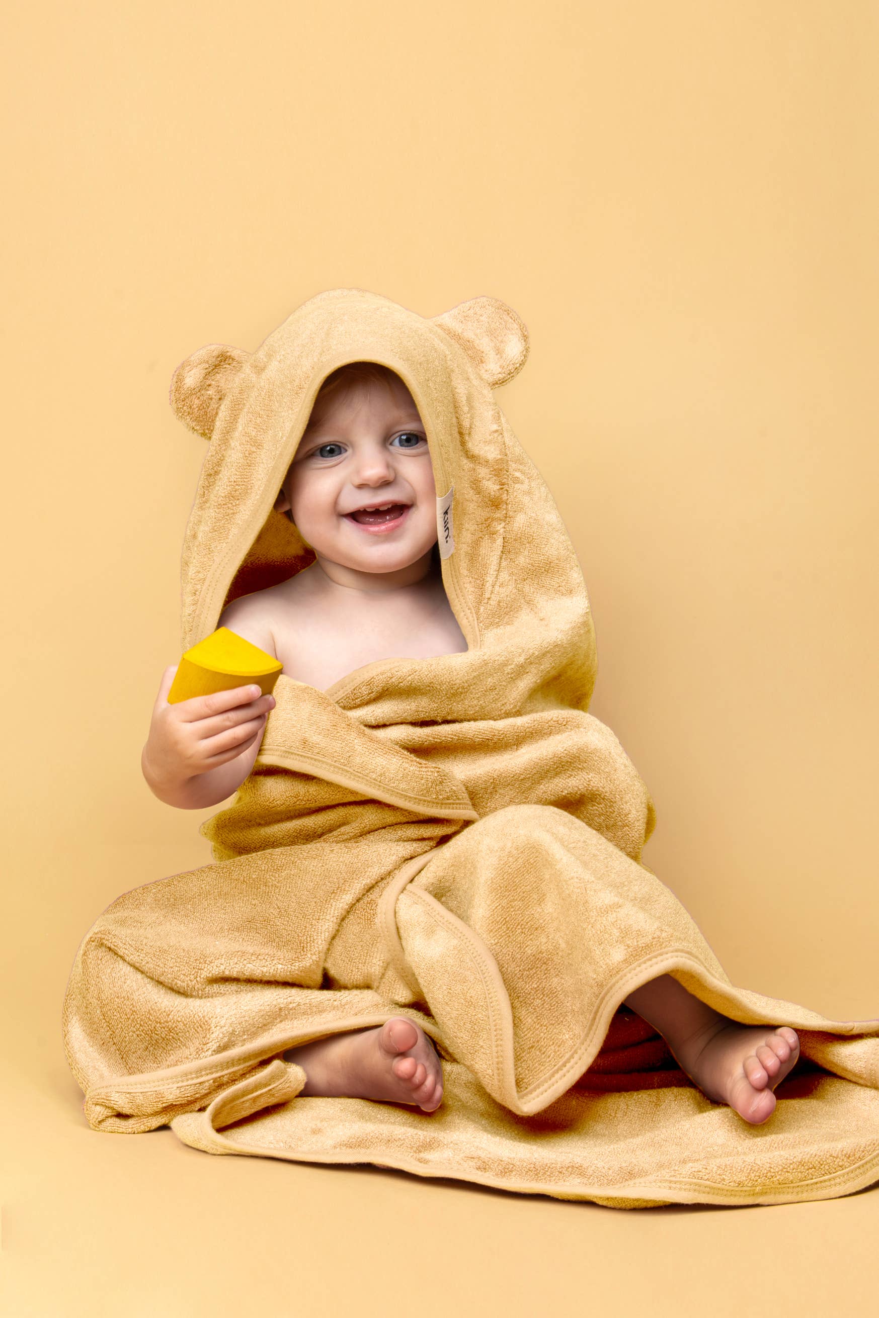 A baby in a KIIN BABY Hooded Towel Ivory, made of hypoallergenic bamboo, smiles on a soft yellow background while holding a yellow block. The towel with bear ears creates a cute and cozy scene.