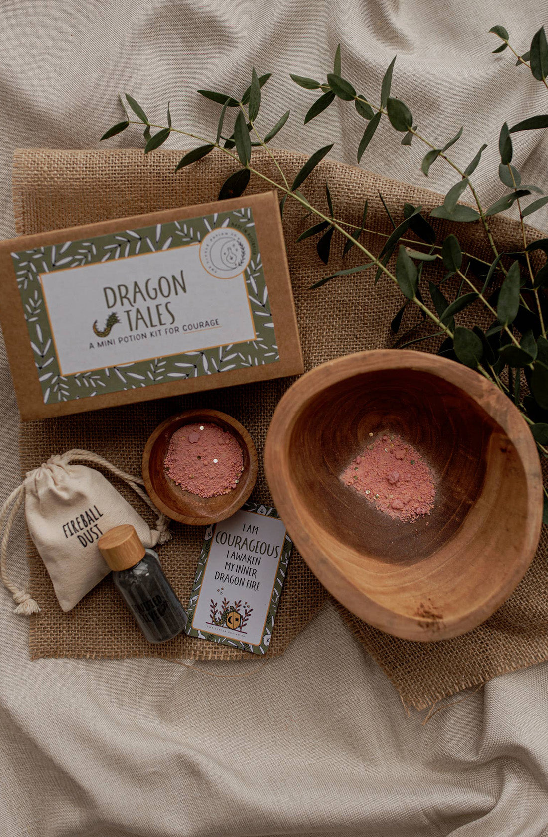 A MINI Dragon Tales Potion Kit (a potion for courage) by THE LITTLE POTION CO is displayed, perfect for imaginative play. The kit includes a glass vial with green contents, a small sack labeled "Fleabane Dust," a wood bowl and dish with pink powder, and a pamphlet titled "Courageous Dragon Fire." Green leaves are scattered around.