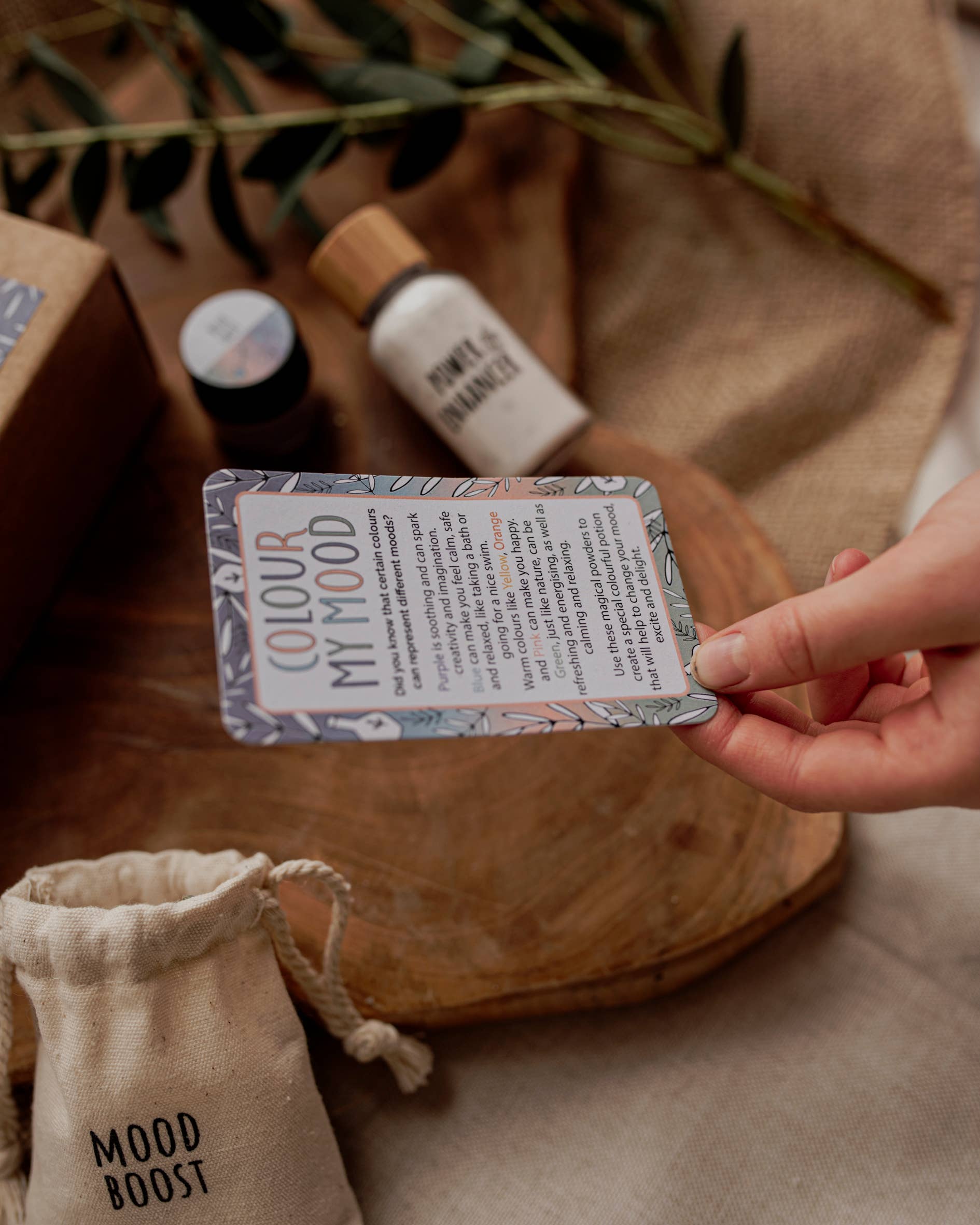 A hand holds a card titled "MINI Colour Mood Potion Kit" by THE LITTLE POTION CO, detailing a mood-boosting activity. Nearby are small bottles, a cloth bag labeled "Mood Boost," and a wooden tray placed on brown textured fabric with green leaves in the background, evoking the essence of sensory play or perhaps magic potion kits.