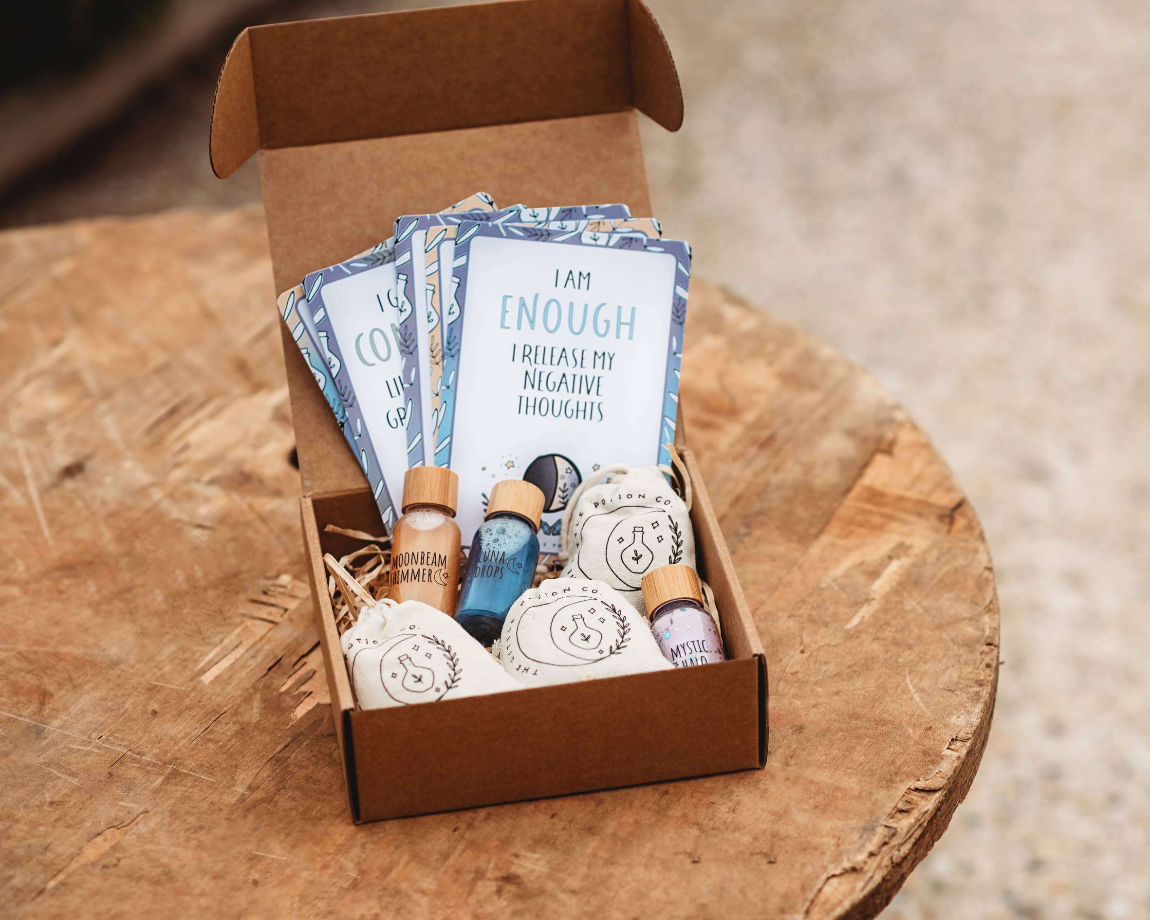 An open cardboard box on a wooden surface contains blue and white cards with positive affirmations, essential oil bottles, and small pouches. The card on top reads, "I AM ENOUGH. I RELEASE MY NEGATIVE THOUGHTS." This Moon Magic MINDFUL Magic Potion Kit by THE LITTLE POTION CO serves as your daily potion kit for inner peace.