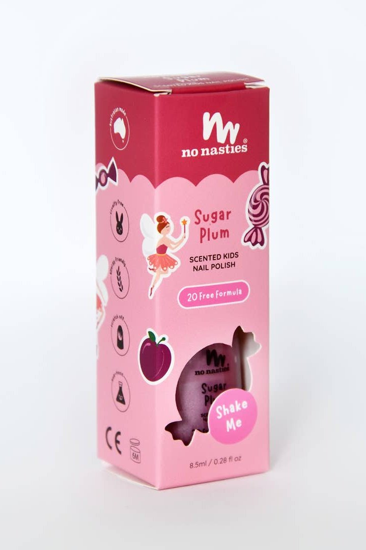 The image shows a pink box containing "Sugar Plum Water Based Scented Scratch Off Kids Nail Polish" by NO NASTIES. It features whimsical illustrations of a fairy, plum, and candy and is proudly labeled with a "20 Free Formula," ensuring it is vegan and cruelty-free. A "Shake Me" note accompanies the cut-out that reveals the non-toxic bottle inside.