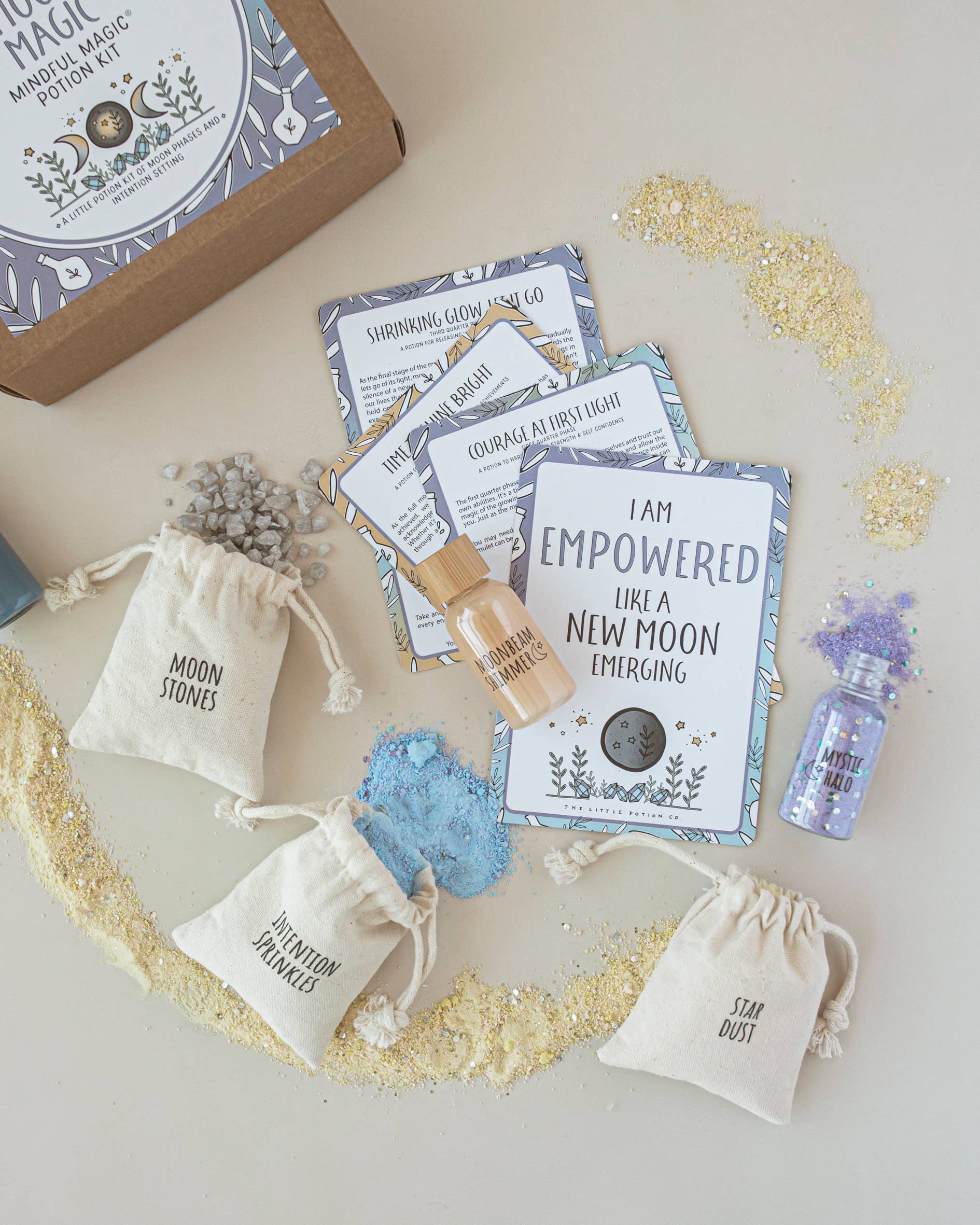 A flat lay of the Moon Magic MINDFUL Magic Potion Kit by THE LITTLE POTION CO makes for the perfect mindfulness activity, featuring three small fabric bags labeled "MOON STONES," "INTENTION STONES," and "STAR DUST." Also shown are cards with affirmations, a small bottle of essential oils, and glittery powders spread around, ideal for any moon phase ritual.