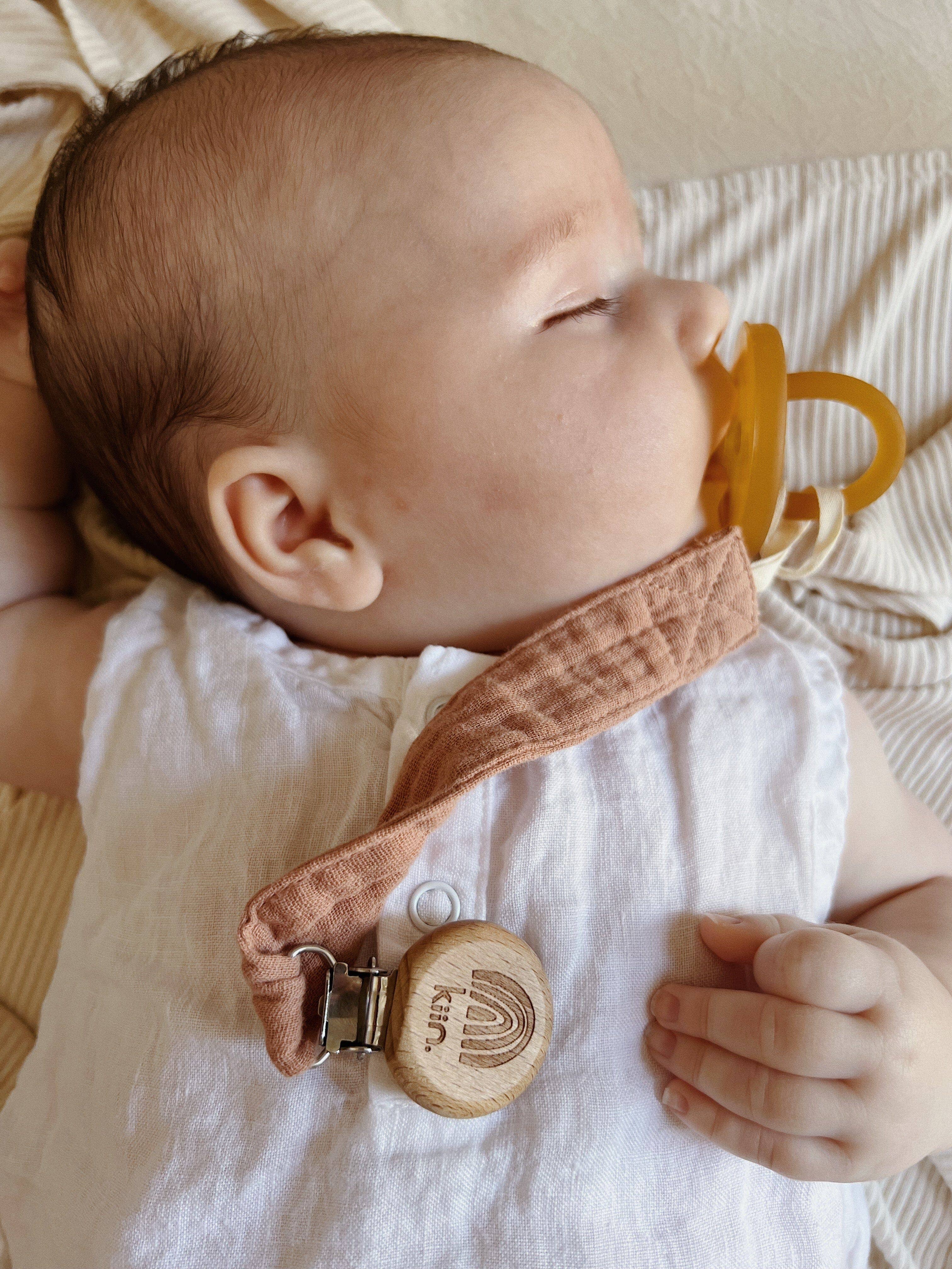 A sleeping baby with a pacifier rests on a beige pillow, wearing a white outfit and the KIIN BABY Cotton Dummy Clip Oat with a wooden clasp, making it perfect for any gender-neutral nursery or as a thoughtful baby shower gift.