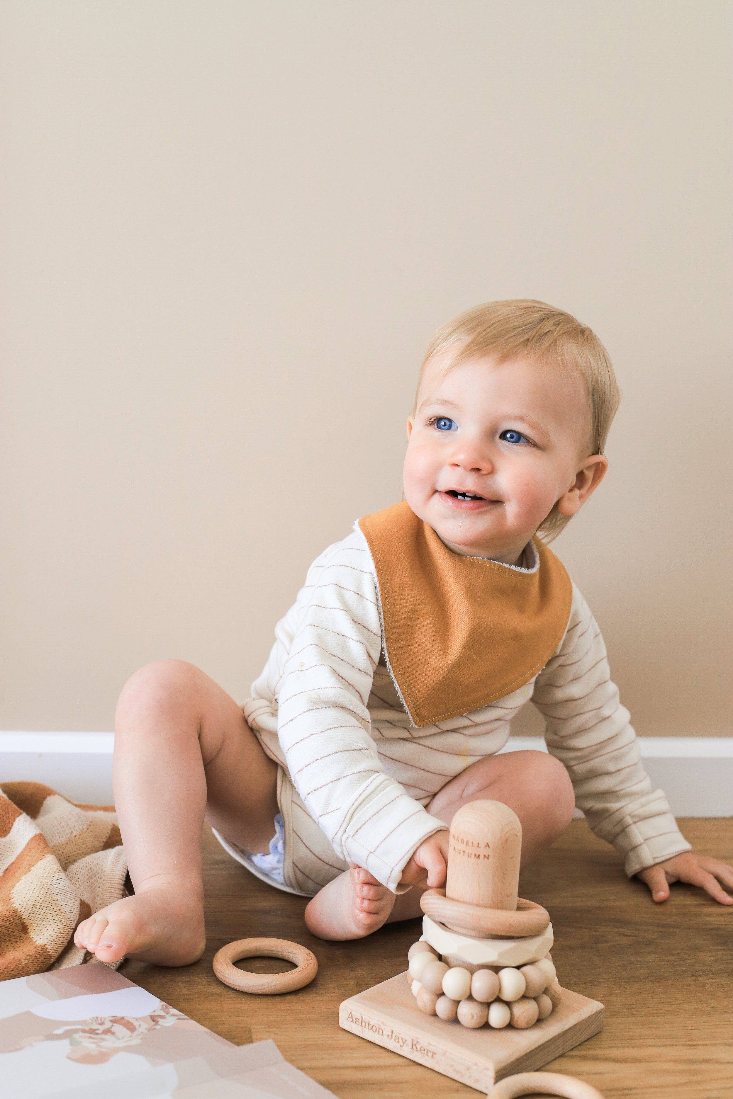 A smiling blonde baby sits on the floor in a striped onesie and Kiin Baby Dribble Bib, surrounded by wooden toys like a ring stacker and rattle. Nearby, a beige blanket and a piece of paper add to the charming scene.