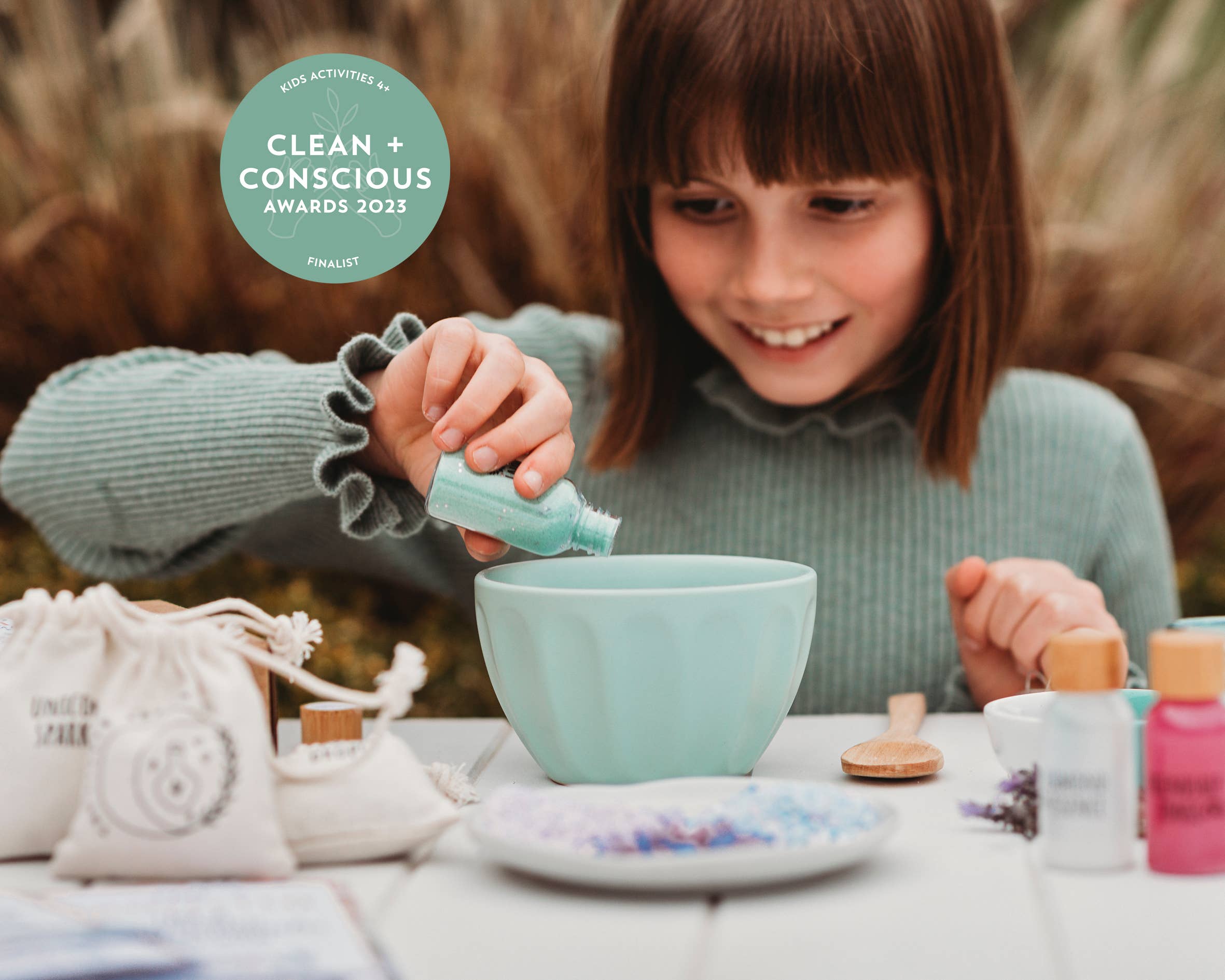 A young girl pours glitter into a light blue bowl while crafting her Rainbow Sparkles MINDFUL Potion Kit by THE LITTLE POTION CO. In front of her are various crafting supplies, including more glitter, a small wooden spoon, and cloth pouches. The image boasts a "Clean + Conscious Awards 2023 Finalist" badge in the top left corner.