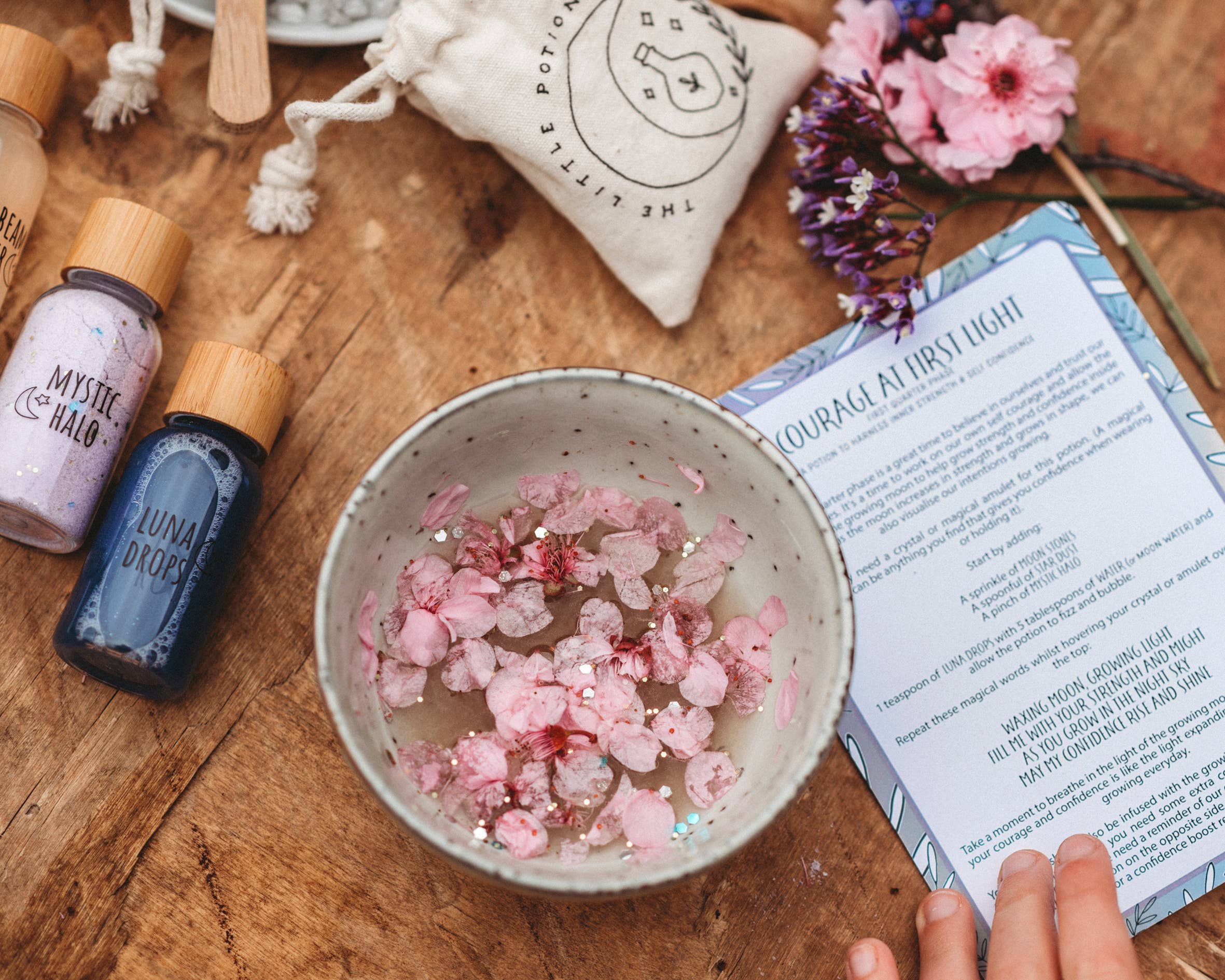 A wooden table displays a bowl filled with pink petals and crystals, accompanied by two bottles labeled "Mystic Halo" and "Luna Drops," some dried flowers, a white drawstring pouch, and a hand holding a sheet titled "Courage at First Light." This thoughtfully arranged scene resembles the Moon Magic MINDFUL Magic Potion Kit by THE LITTLE POTION CO, inspired by moon phases.