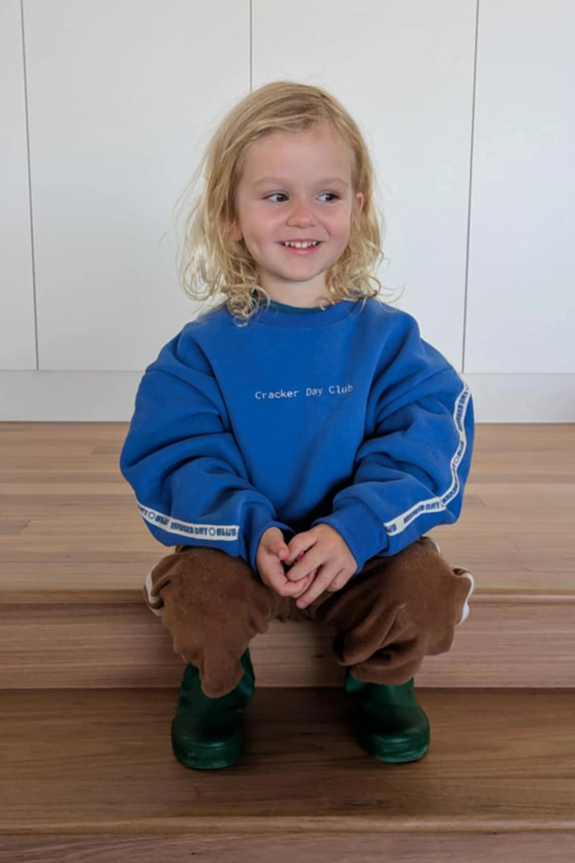 A young child with long blond hair, wearing an oversized Cracker Day Club - Kids Cloud Racer Sweater Blue by CRACKER DAY CLUB, brown pants, and green rain boots, sits on wooden steps smiling and looking slightly to the side.