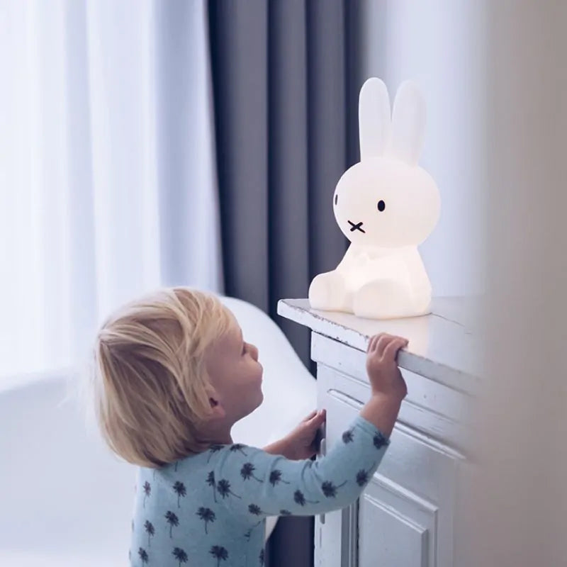 A young child with light blonde hair, wearing a light blue long-sleeve shirt adorned with small tree prints, gazes curiously at the glowing white MR MARIA Miffy First Light Lamp placed on a white dresser. The room features neutral tones with soft lighting and a curtained window in the background.