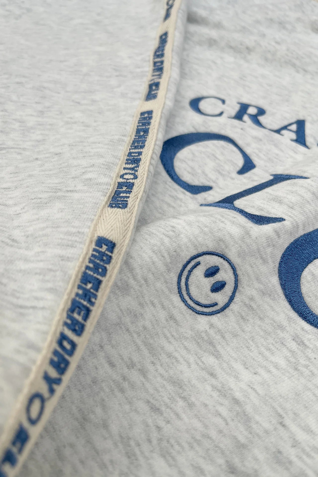 A close-up of heavyweight terry cotton fabric with blue text and a smiley face. A beige seam with repeating blue "CRACKER DAY CLUB" text runs diagonally across the Cloud Quarter Zip Sweater Grey Marle.