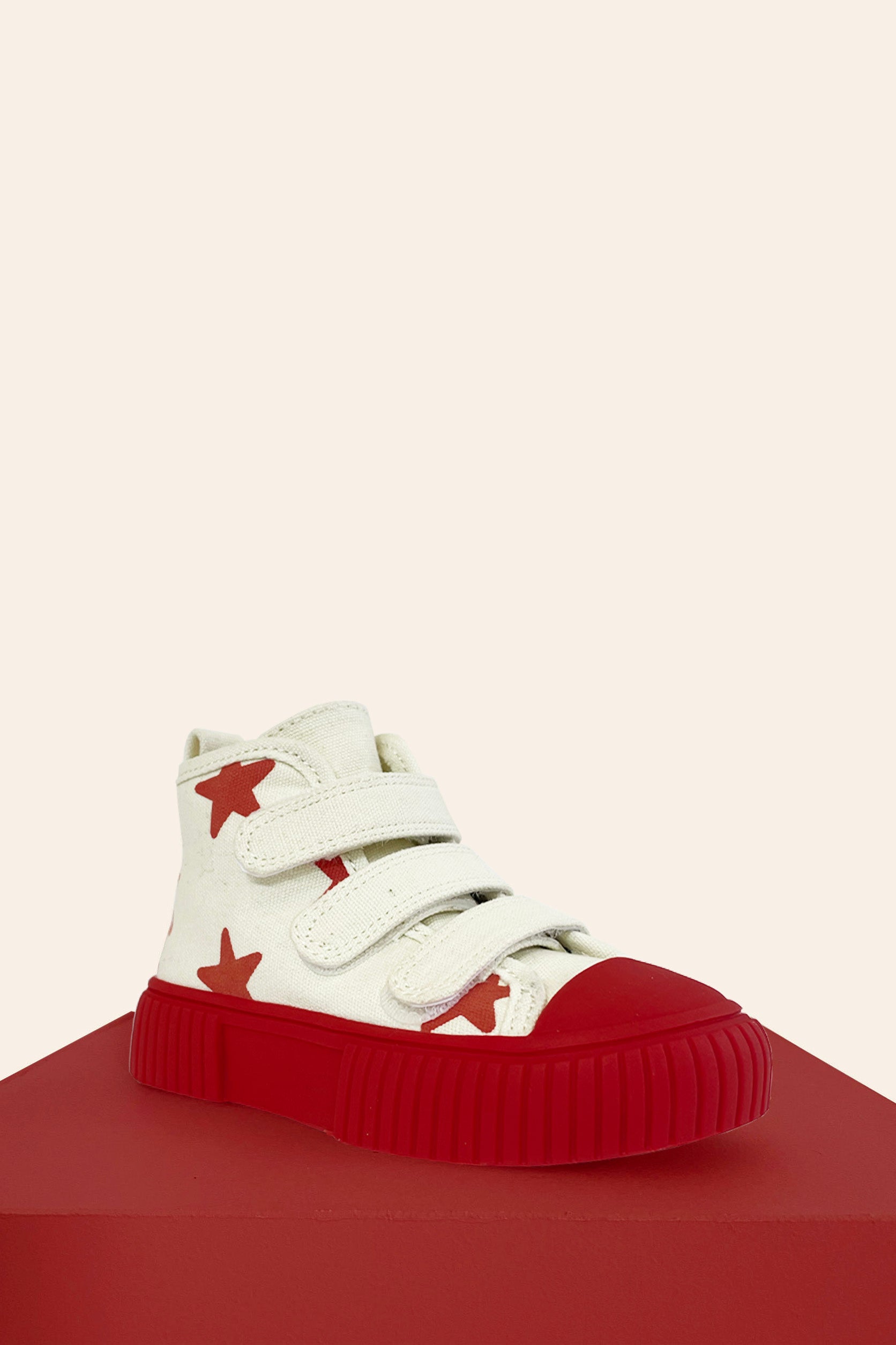 The PRE-ORDER Limited Edition Piccolini x By Billie High Top Sneaker Red Stars by PICCOLINI is a children's shoe designed with white uppers embellished with red star patterns and made from organic cotton. It features three Velcro straps and comes with a podiatrist-recommended, non-slip rubber sole. The shoe is showcased on a red platform against a beige background.