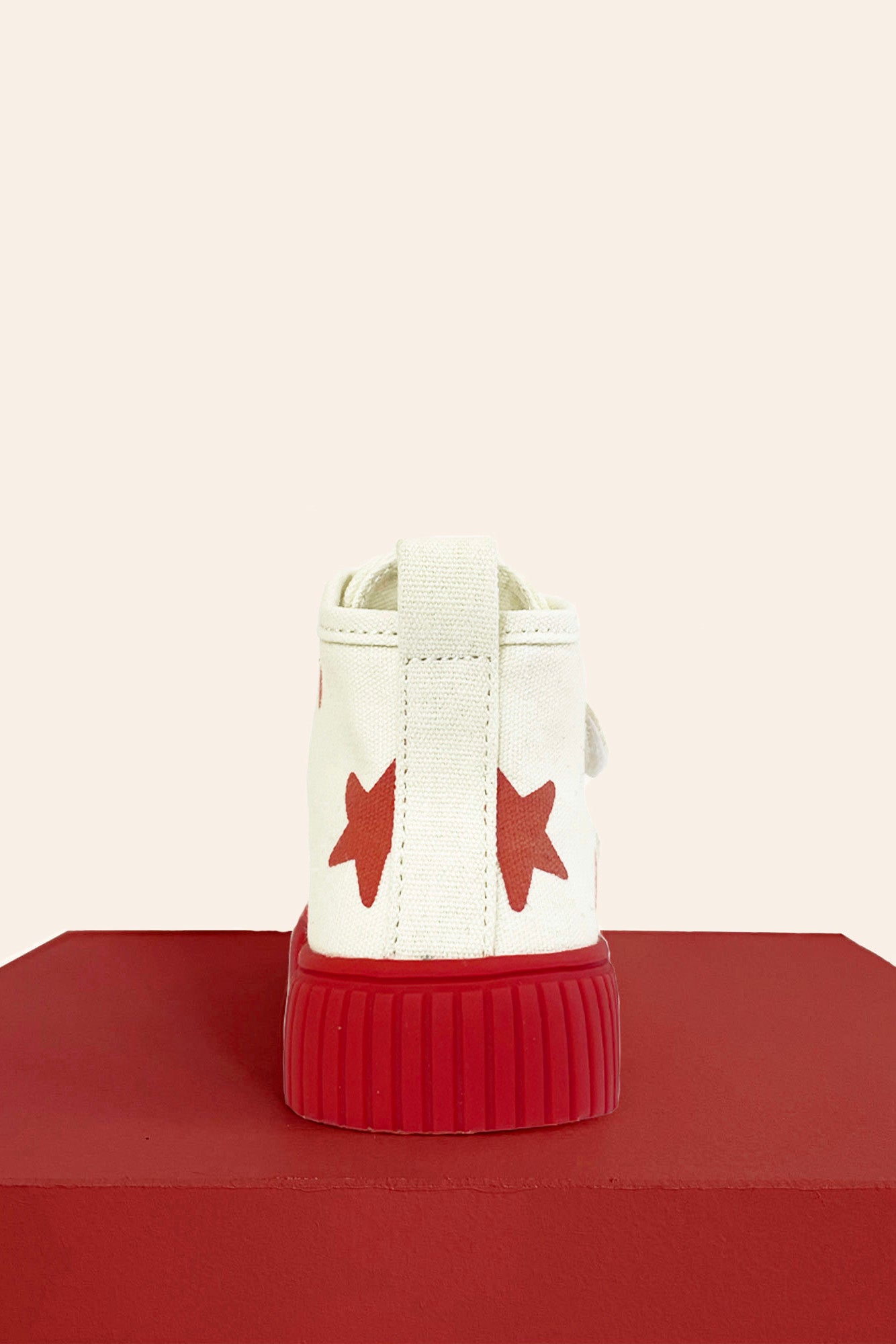 The PRE-ORDER Limited Edition Piccolini x By Billie High Top Sneaker Red Stars, crafted from organic cotton and adorned with red star patterns on the side, is displayed on a red platform against a light beige background. This PICCOLINI shoe is shown from the back to highlight its non-slip rubber sole and distinctive red sole.