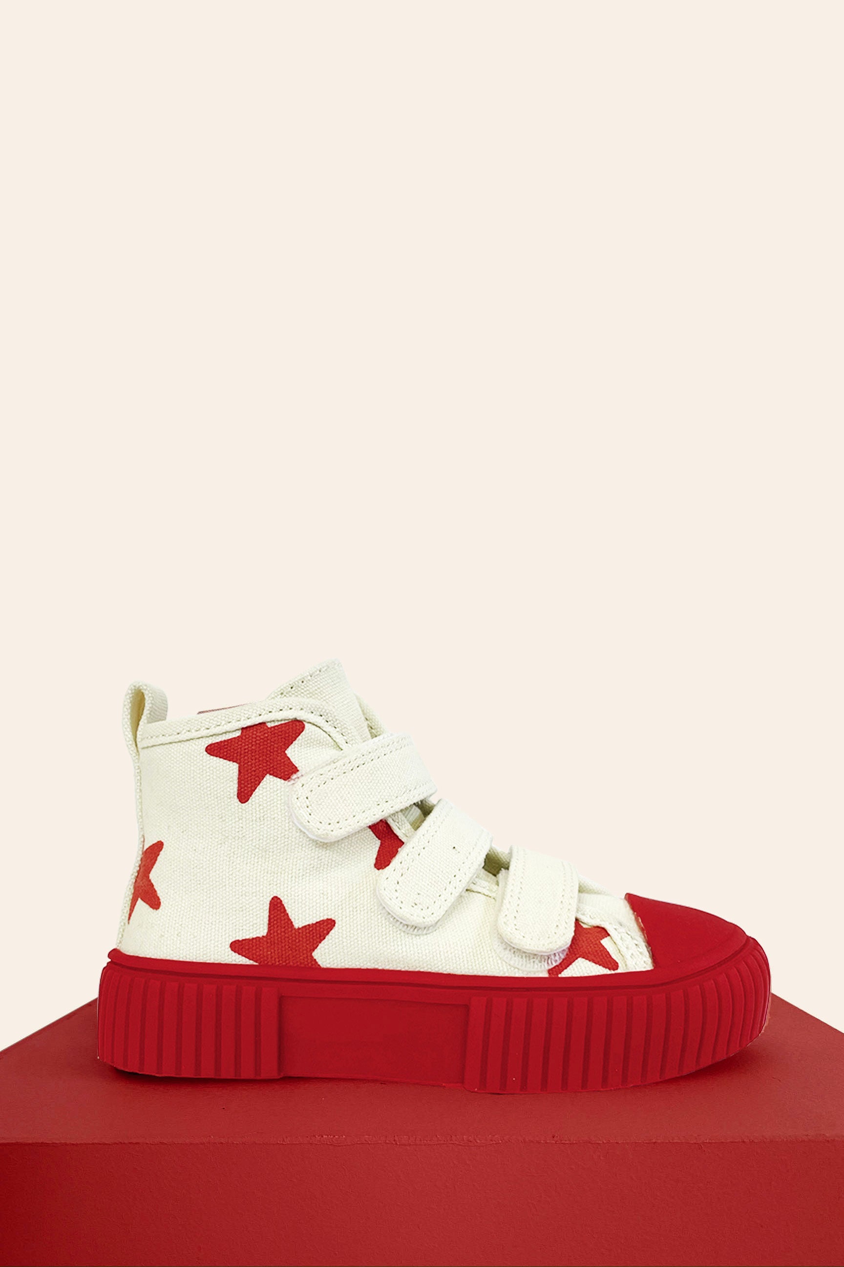 The PRE-ORDER Limited Edition Piccolini x By Billie High Top Sneaker Red Stars by PICCOLINI is a high-top sneaker made from podiatrist-recommended organic cotton. It features a white canvas body adorned with red stars and equipped with durable, non-slip rubber soles. The design includes three white Velcro straps and is presented on a red platform against a beige background.