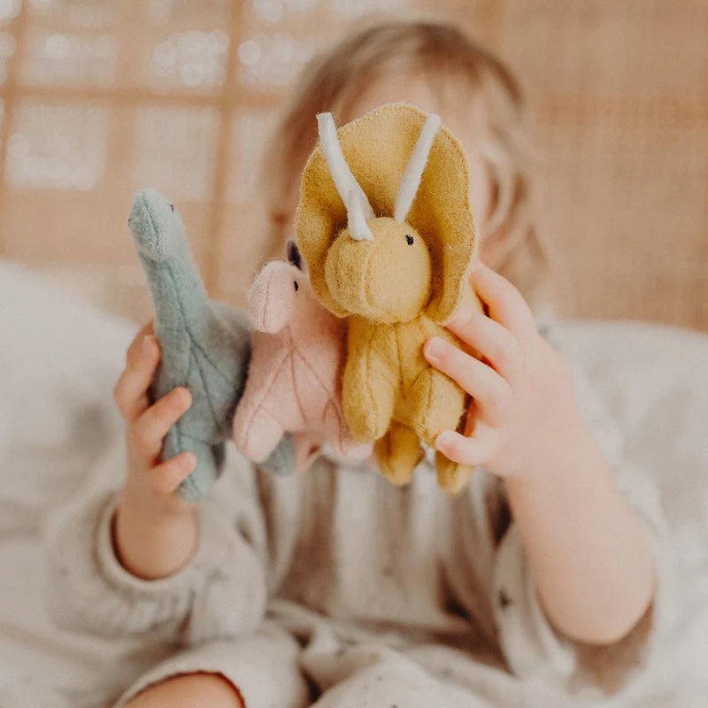 A small child, mostly out of focus, holds three colorful OLLI ELLA Holdie Dinosaurs: a green one, a pink one, and a yellow one. They are sitting on a white bedspread with a soft background suggesting a cozy room filled with prehistoric pals.