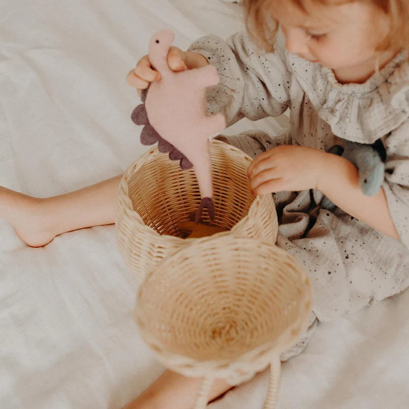 A young child sits on a white surface, wearing an outfit made from a handmade soft wool blend, and places their OLLI ELLA Holdie Dinosaur, a pink plush toy, into a woven basket. The vibrant dinosaur stands out beautifully against the grey of their attire. Nearby, there's a second smaller basket.