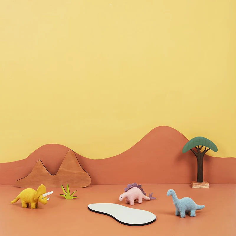 A vibrant Holdie Dinosaurs diorama from OLLI ELLA features three toy dinosaurs: a yellow triceratops, a pink stegosaurus, and a blue brachiosaurus. These prehistoric friends gather around a small white pond set against an orange and yellow landscape adorned with mountains, a plant, and a tree in the background.