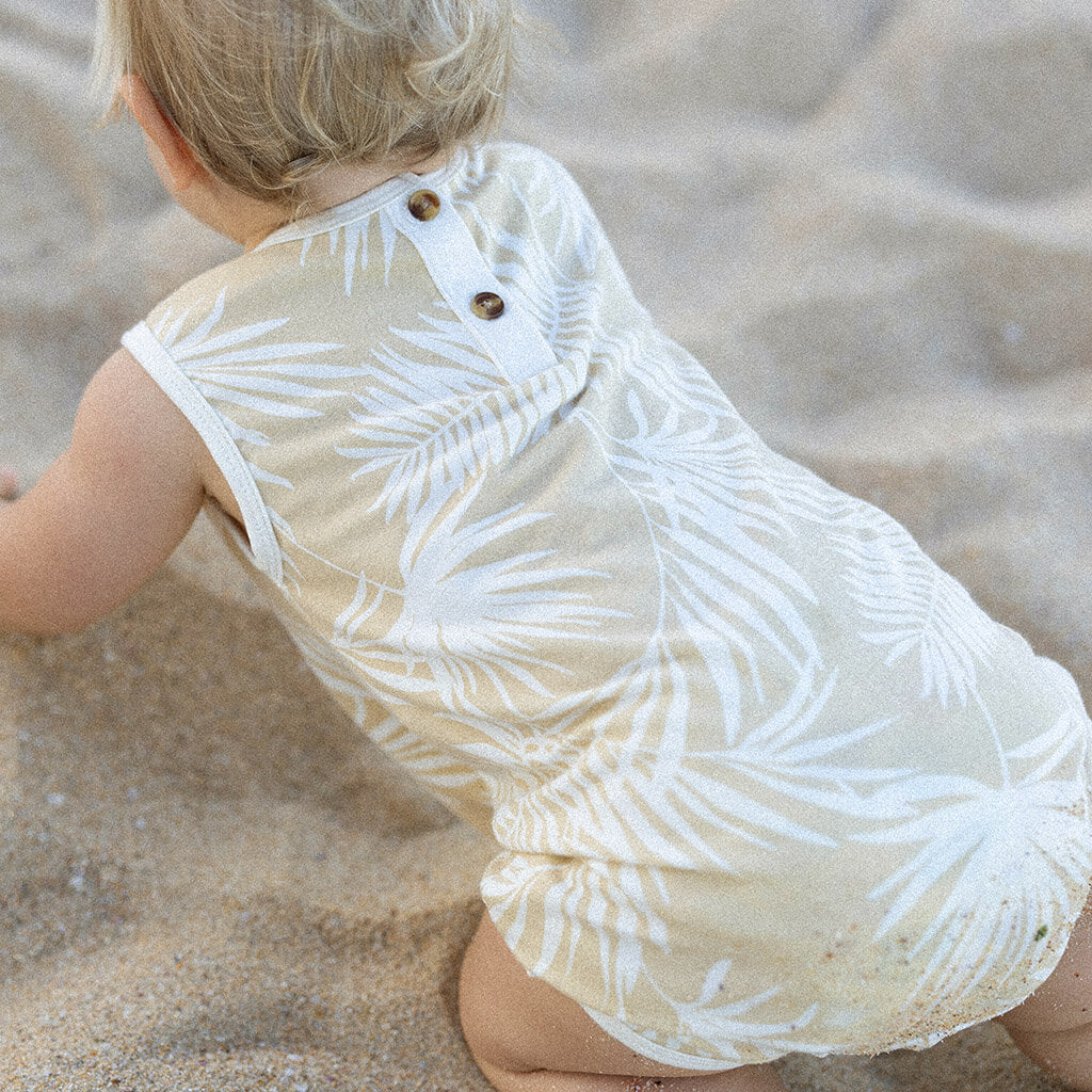 A toddler in a sleeveless, light-colored Hula Playsuit by BAM LOVES BOO with a vintage palm print is playing on sandy ground. The child's hair is light and wavy. The focus is on the back of the toddler, highlighting the eco-friendly dyes and button details of their adorable attire.