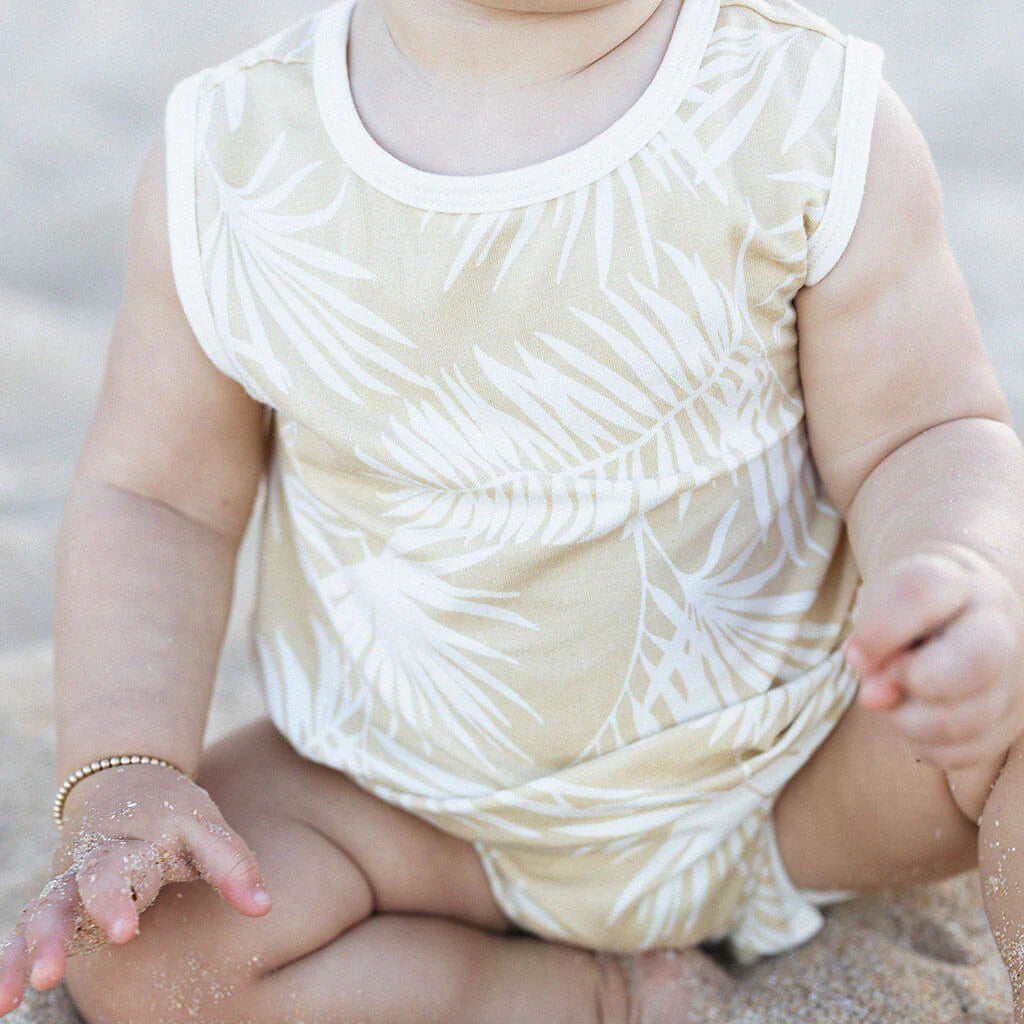 A baby sits on sandy ground wearing the Hula Playsuit by BAM LOVES BOO, a light-colored, sleeveless outfit featuring a vintage palm print. The baby's hand touches the sand, highlighting the super soft 2-way stretch fabric that ensures comfort and flexibility.