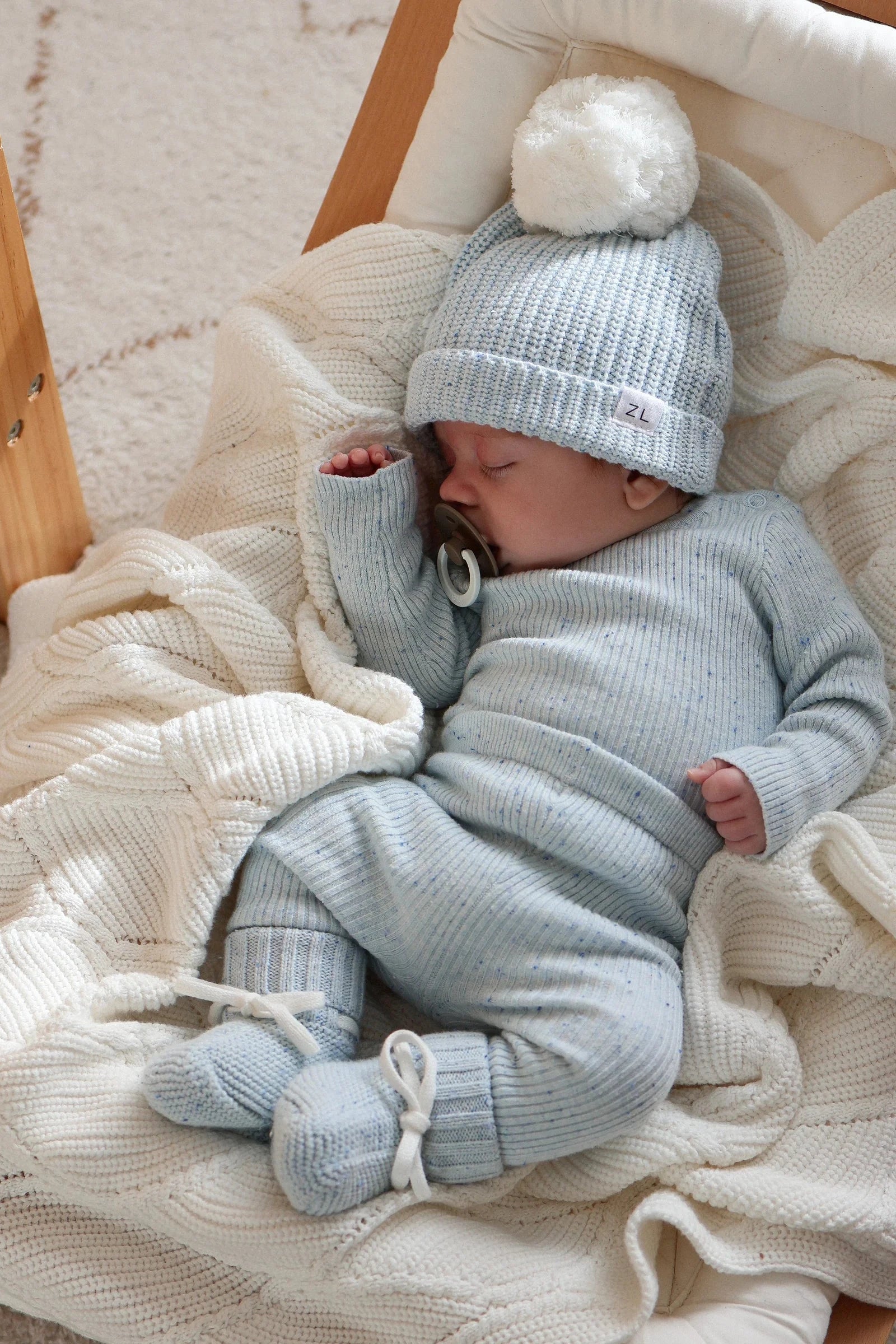 A baby sleeps in a cozy wooden cradle, wrapped in a white knitted blanket. The baby is dressed in light blue ribbed pajamas with matching socks and a knit hat adorned with a white pom-pom. Nearby, ZIGGY LOU Booties Aero Fleck sit beside the pacifier resting by the baby's hand.