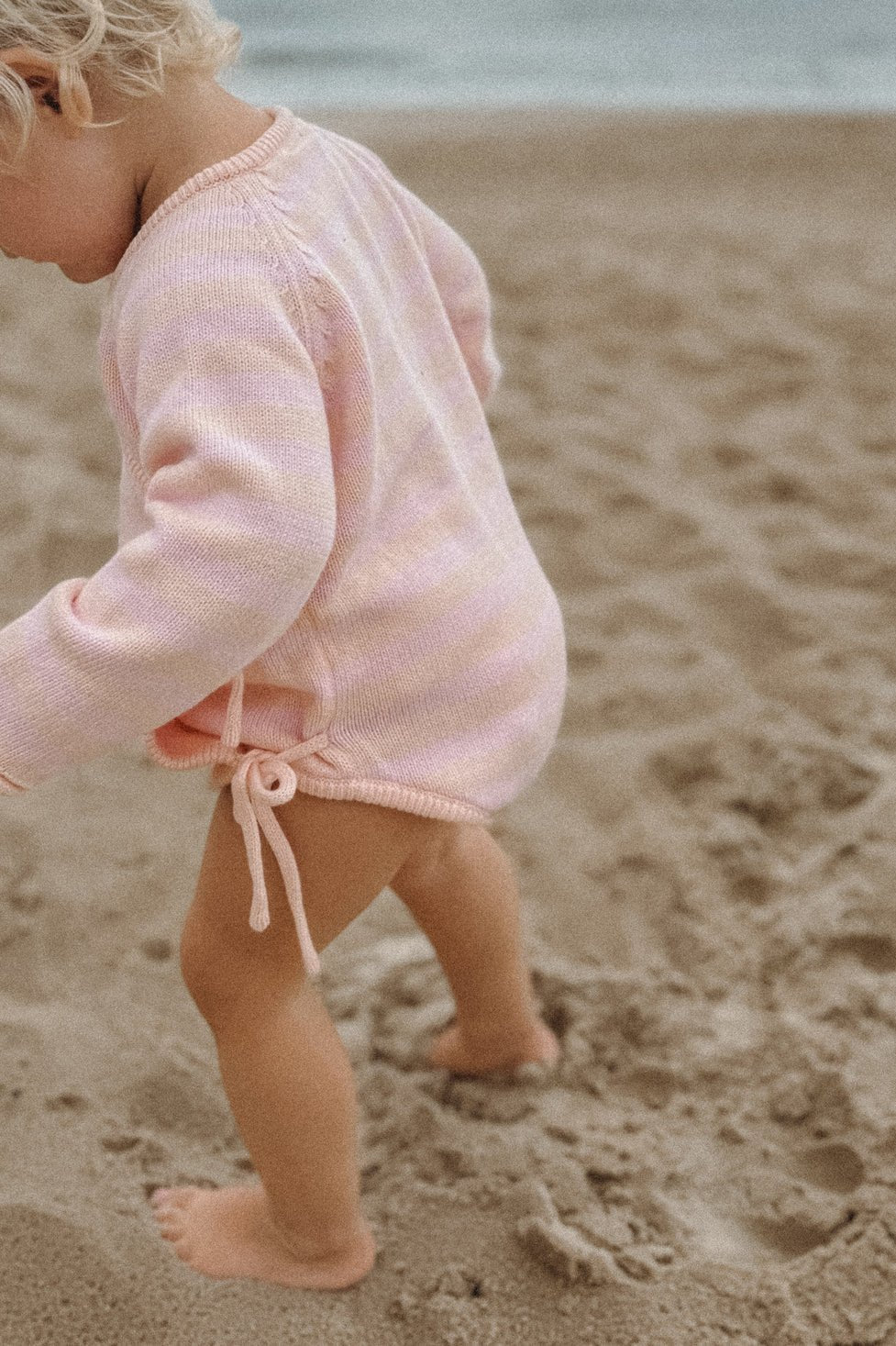 A toddler with curly blonde hair, wearing MILKY DESIGNS' comfortable Essential Wrap Romper in pink/peach, walks barefoot on a sandy beach.