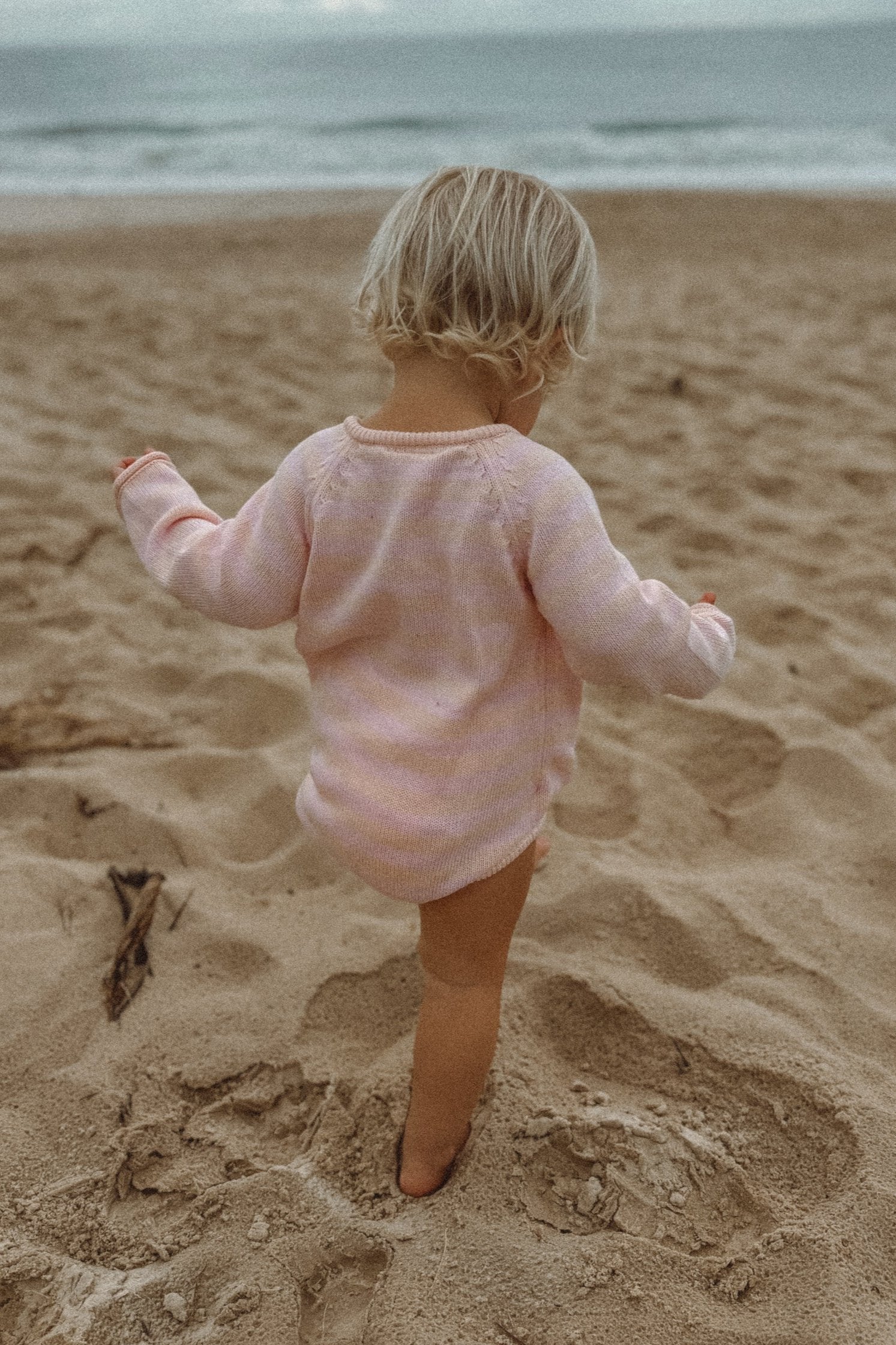 A young child stands with arms slightly outstretched and faces away from the camera on a sandy beach. Wearing the Essential Wrap Romper Pink/Peach from MILKY DESIGNS, crafted from 100% cotton, the child has light blonde hair. The comfort of MILKY DESIGNS baby clothing is evident as the ocean stretches in the background under an overcast sky.