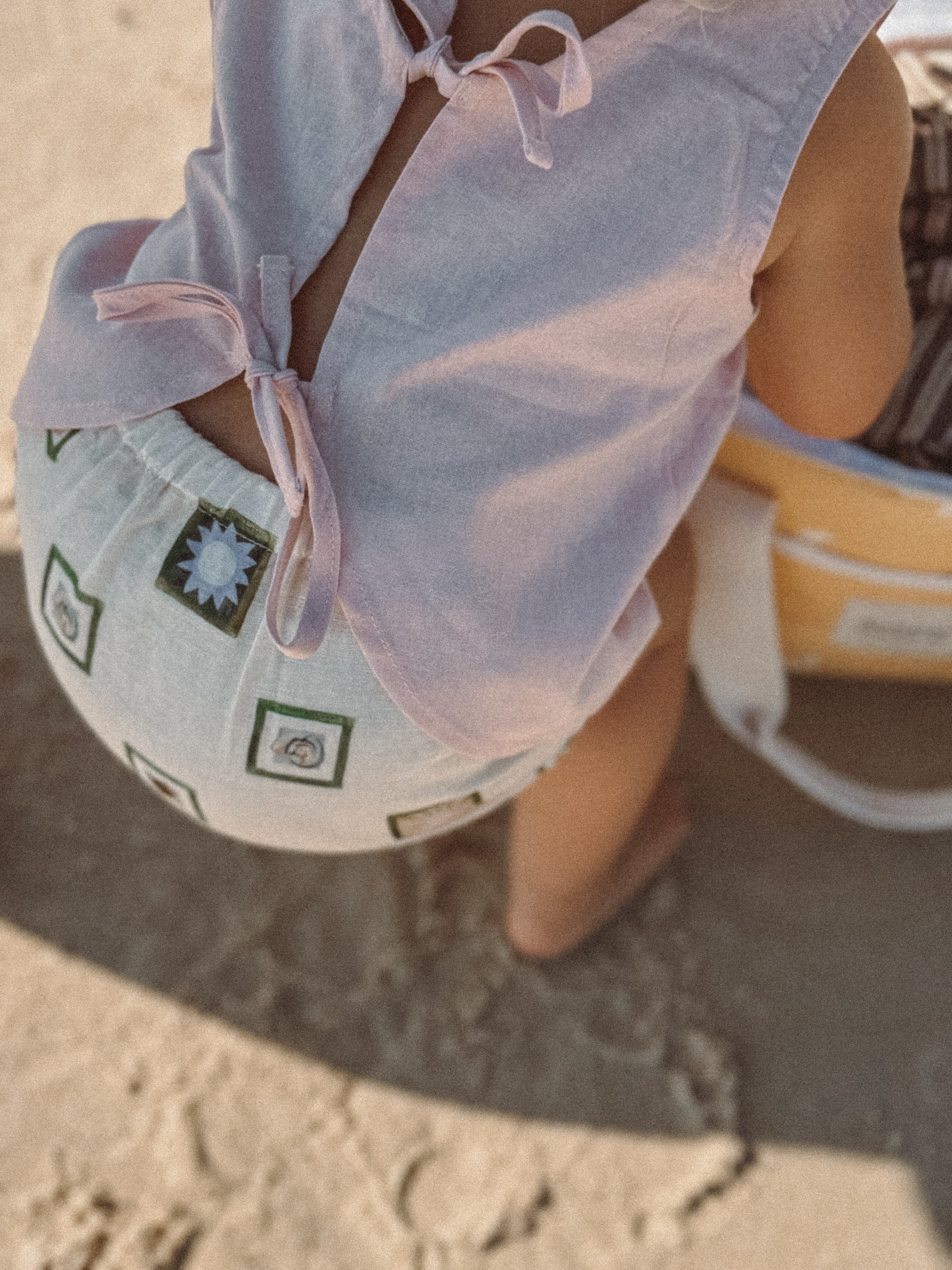 A child in an Annie Bloomers outfit by MILKY DESIGNS, featuring a sleeveless top and patterned shorts with an elastic waist, sits on the sand at the beach. The sun casts a warm glow, illuminating a nearby beach bag.