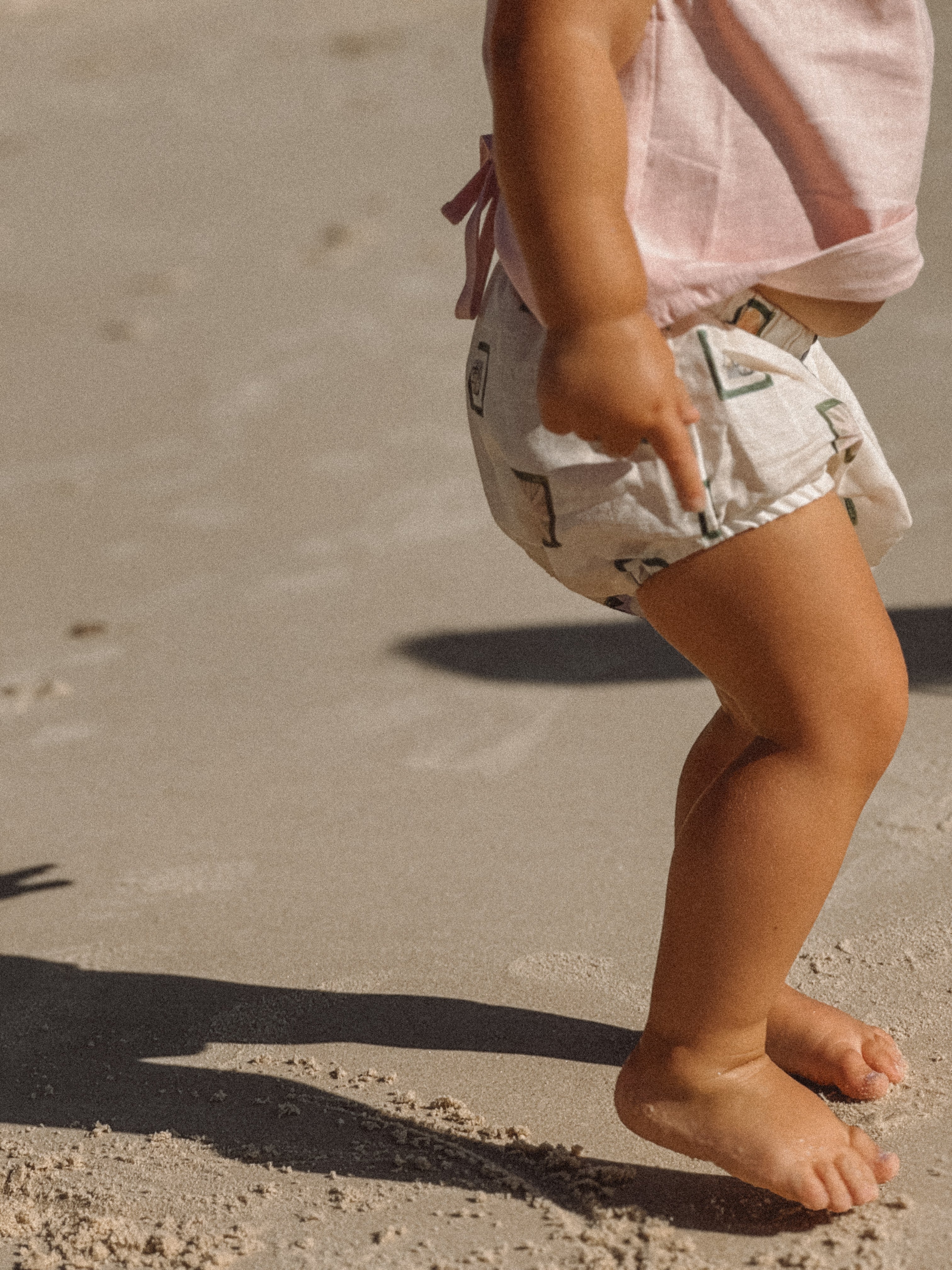 A young child wearing the Annie Bloomers by MILKY DESIGNS, a pink balloon-style top paired with white elastic-waist shorts, stands barefoot on a sandy beach. The child is mid-movement, their shadow dancing alongside them on the sun-kissed sand.