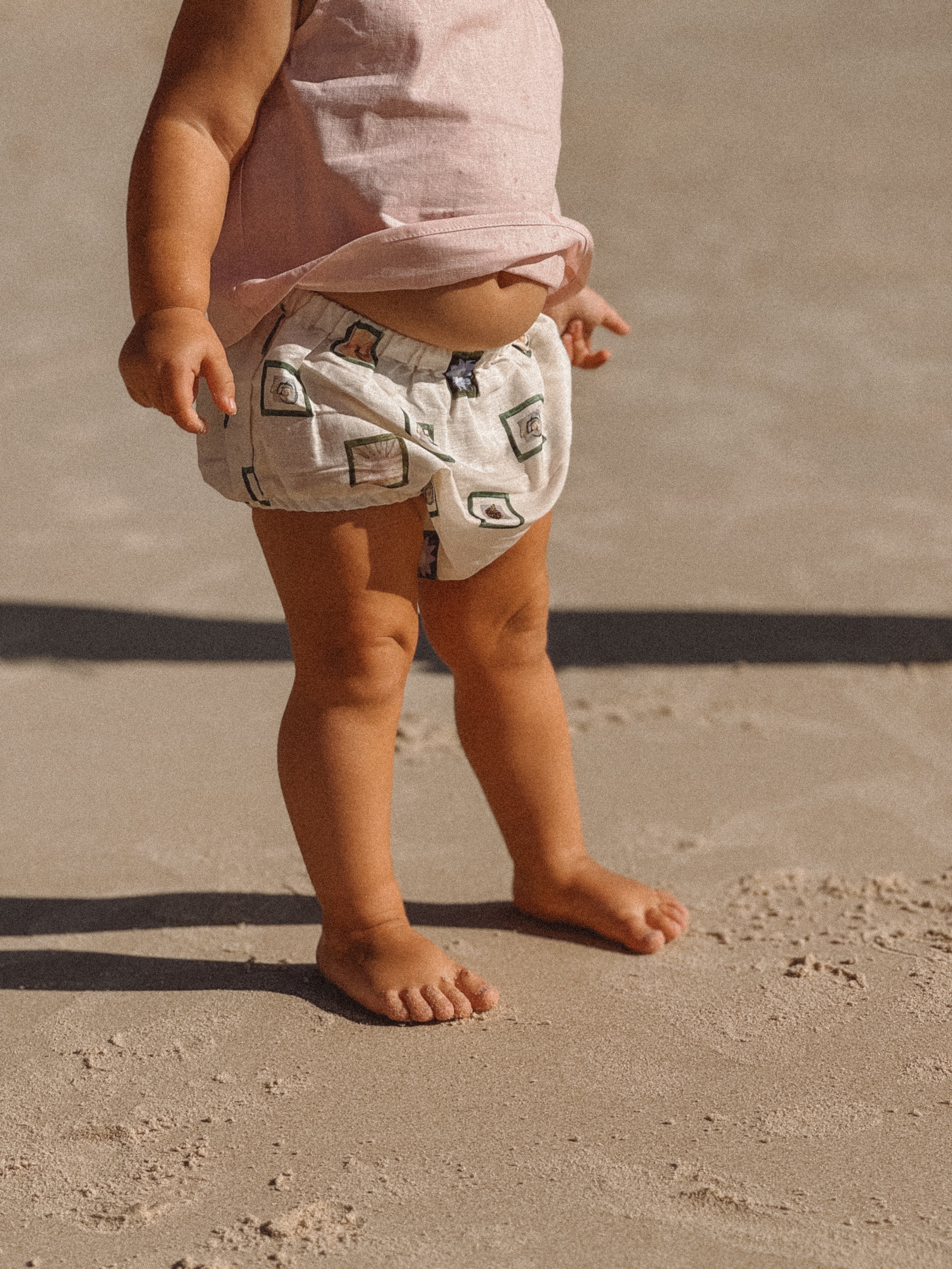 A toddler wearing the Annie Bloomers from MILKY DESIGNS, featuring an elastic waistband and a charming pattern, stands on a sandy beach. The focus is on the child's legs and feet as they touch the sunlit sand, capturing the essence of a warm day at the beach.