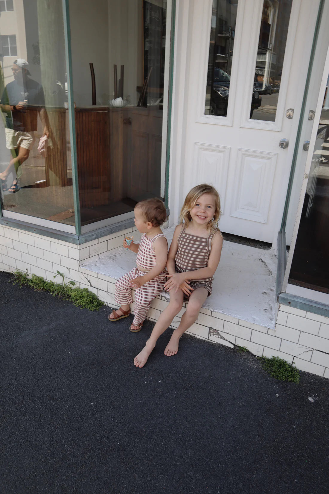 Two young children sit on a step outside a building. The older child, wearing the Bobbi Rib Dress in Choc Stripe from MILKY DESIGNS, smiles at the camera while the younger one looks away, holding a small object. Both wear outfits featuring vibrant colorways. The entrance includes a glass window and a white door.