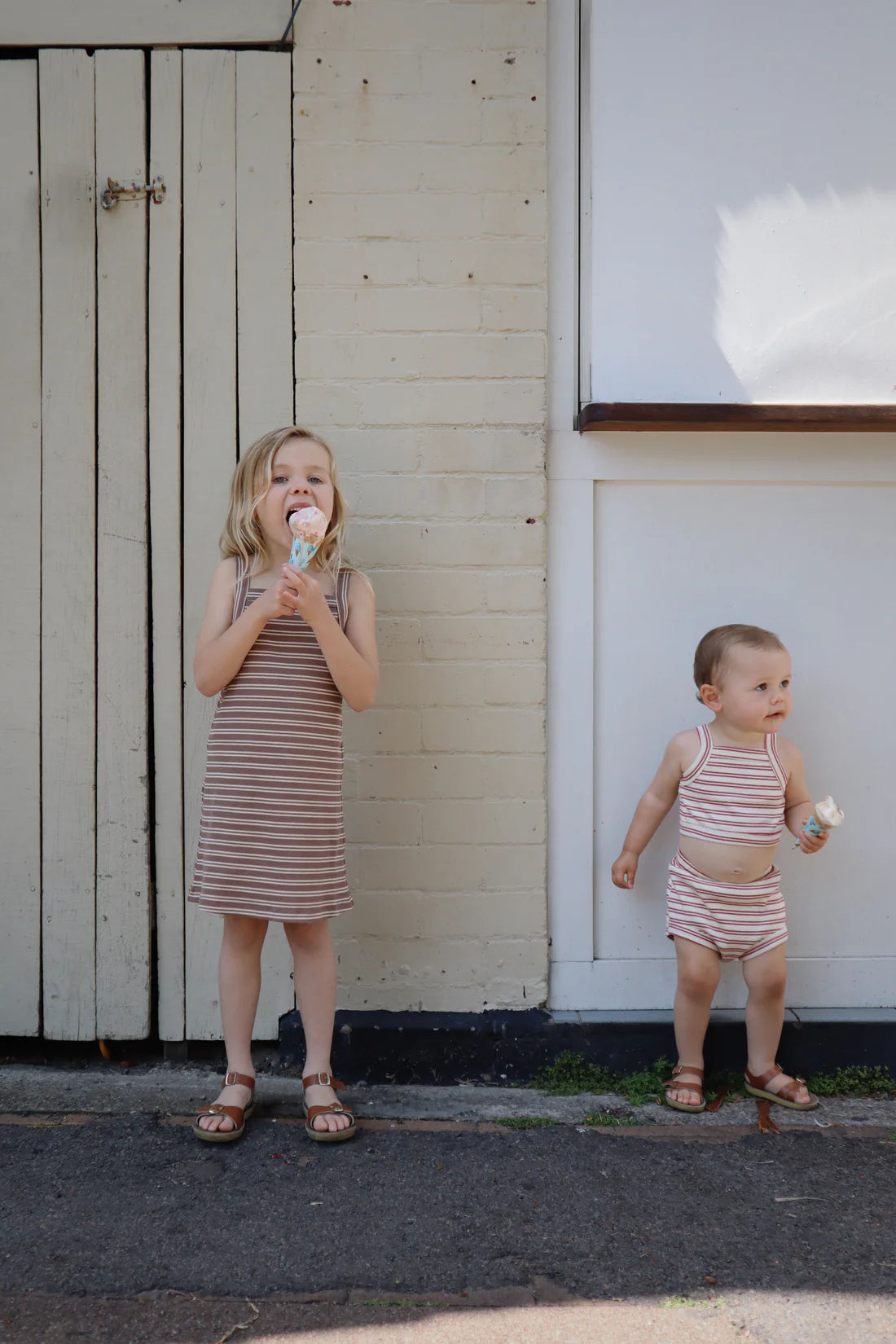 Two young children in matching ribbed cotton outfits stand outside. The older child, wearing a cheerful Bobbi Rib Dress Choc Stripe by MILKY DESIGNS, holds an ice cream cone and smiles, while the younger one also has an ice cream but looks to the side. They are in front of a light-colored wall and door.