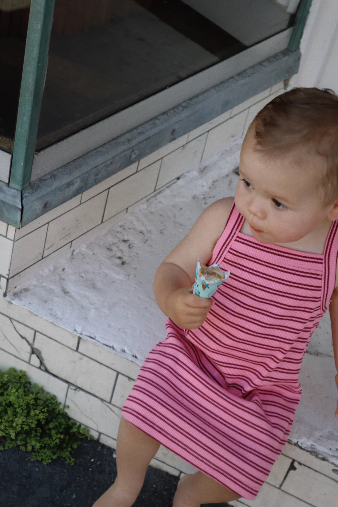 A small child wearing a pink Bobbi Rib Dress with red stripes from MILKY DESIGNS sits on a white step, holding an ice cream cone. The child looks to the right, with a window and greenery visible in the background.